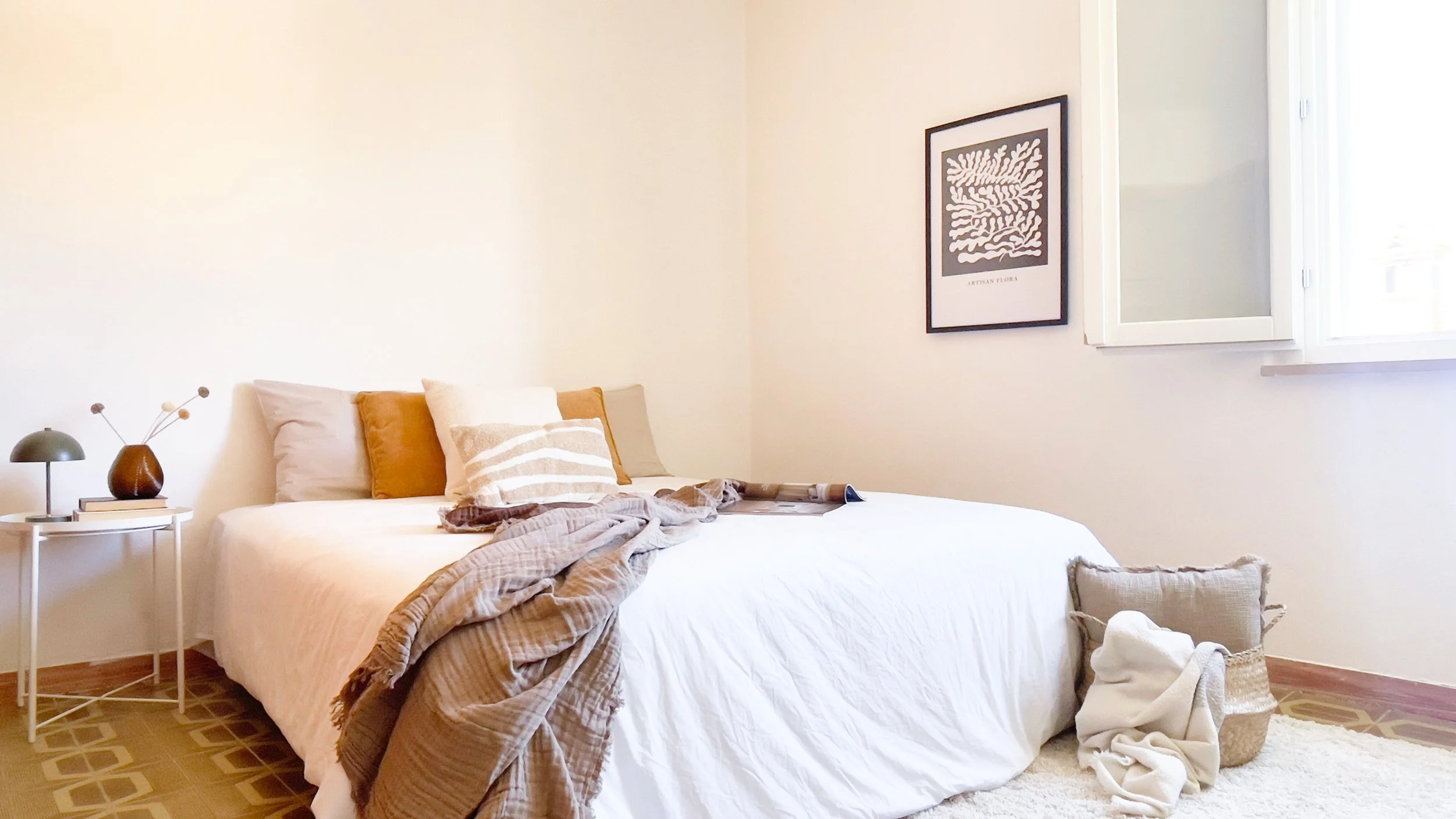Camera da letto minimalista con letto matrimoniale, cuscini, tavolino bianco con lampada e vasi decorativi, finestra e quadro alla parete.