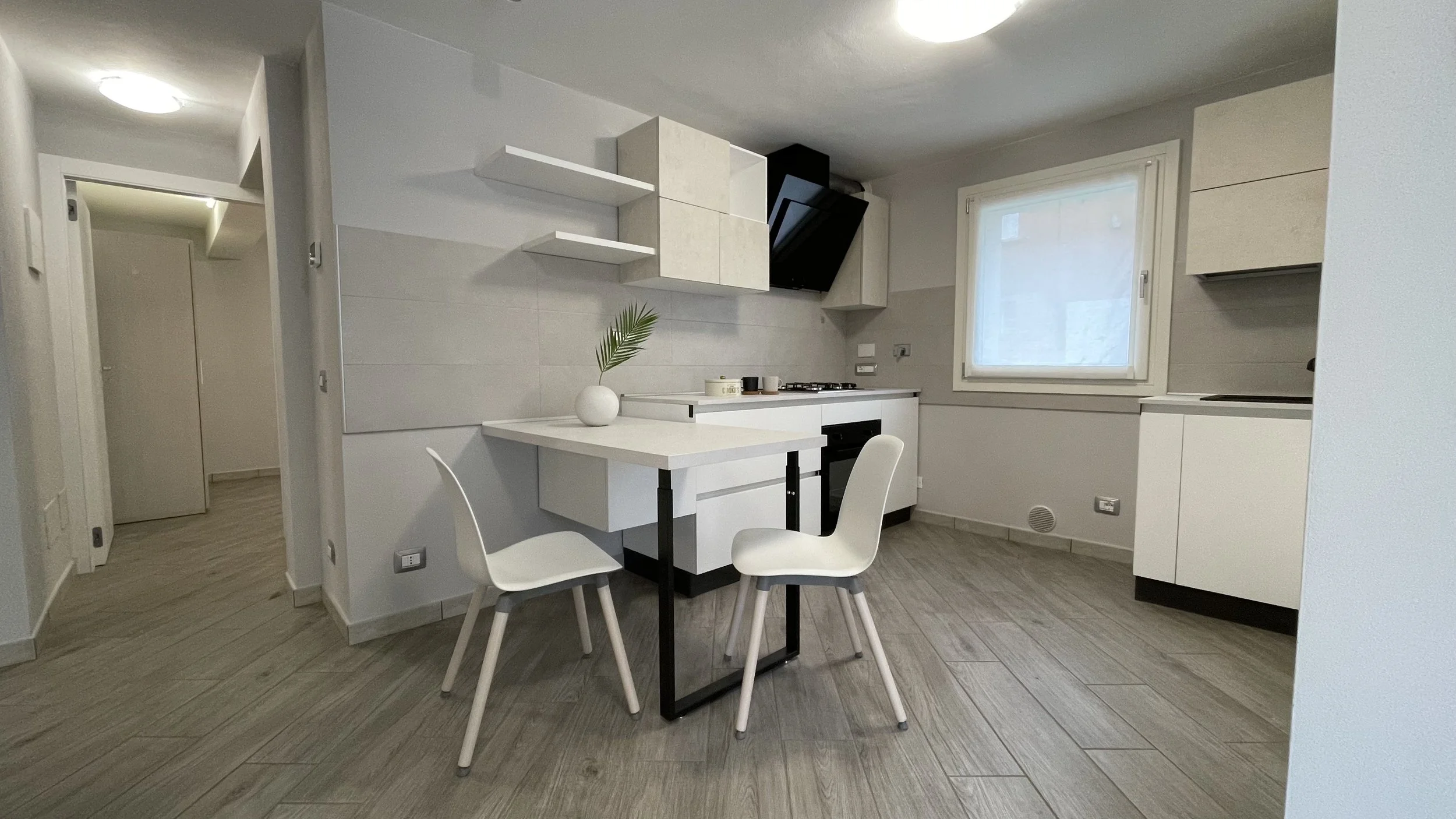 Cucina moderna con tavolo bianco, due sedie, armadietti bianchi e grigi, piano cottura, cappa nera, finestra con tende bianche, e pavimento in legno chiaro.