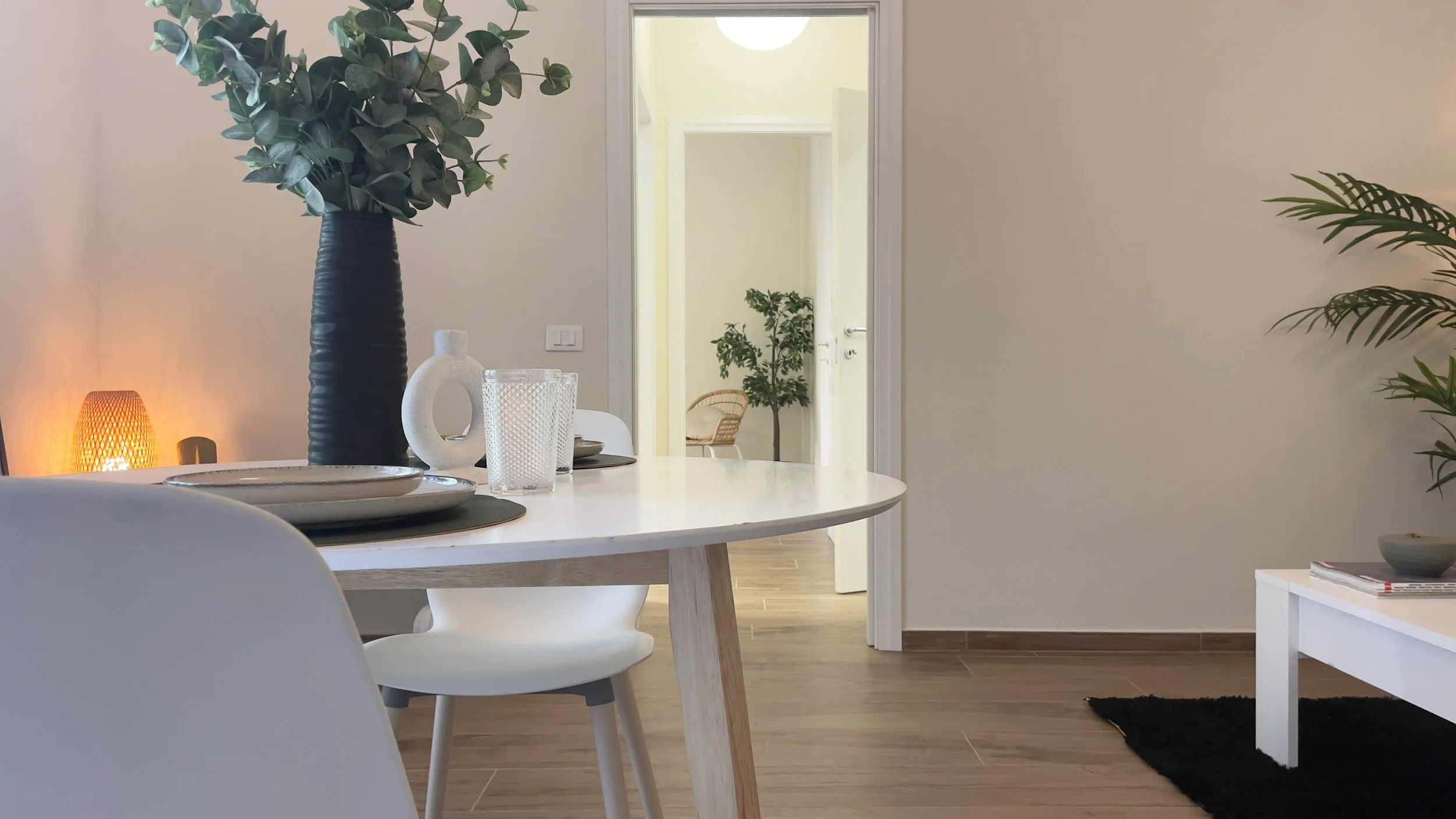 Interno moderno con tavolo bianco, sedie, vaso con foglie e lampada accesa.
