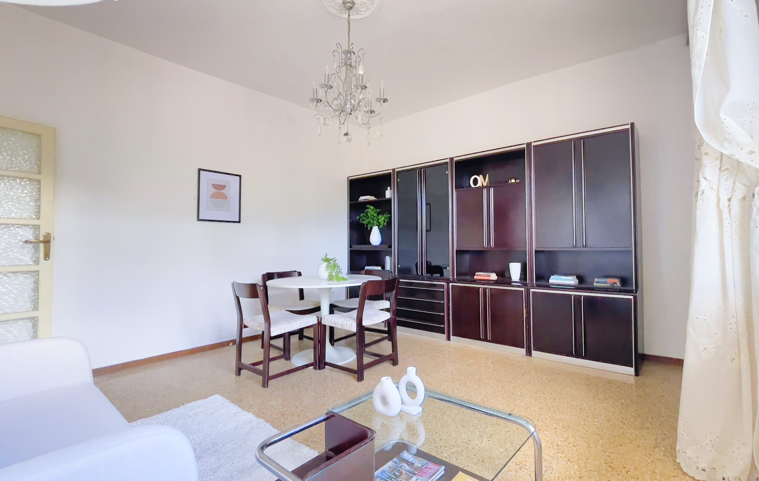 Soggiorno con parete bianca, tavolo da pranzo rotondo bianco con quattro sedie, mobile nero a parete, lampadario a cristallo, decorazioni vegetali e vetrina con riviste.