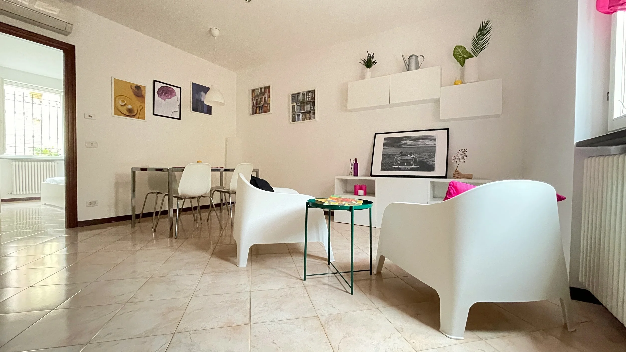 Soggiorno moderno con poltrone bianche, tavolo con sedie bianche, e decorazioni murali. Sullo sfondo, quadri appesi e una mensola con piante decorative. Arredamento semplice e luminoso con pavimento piastrellato.