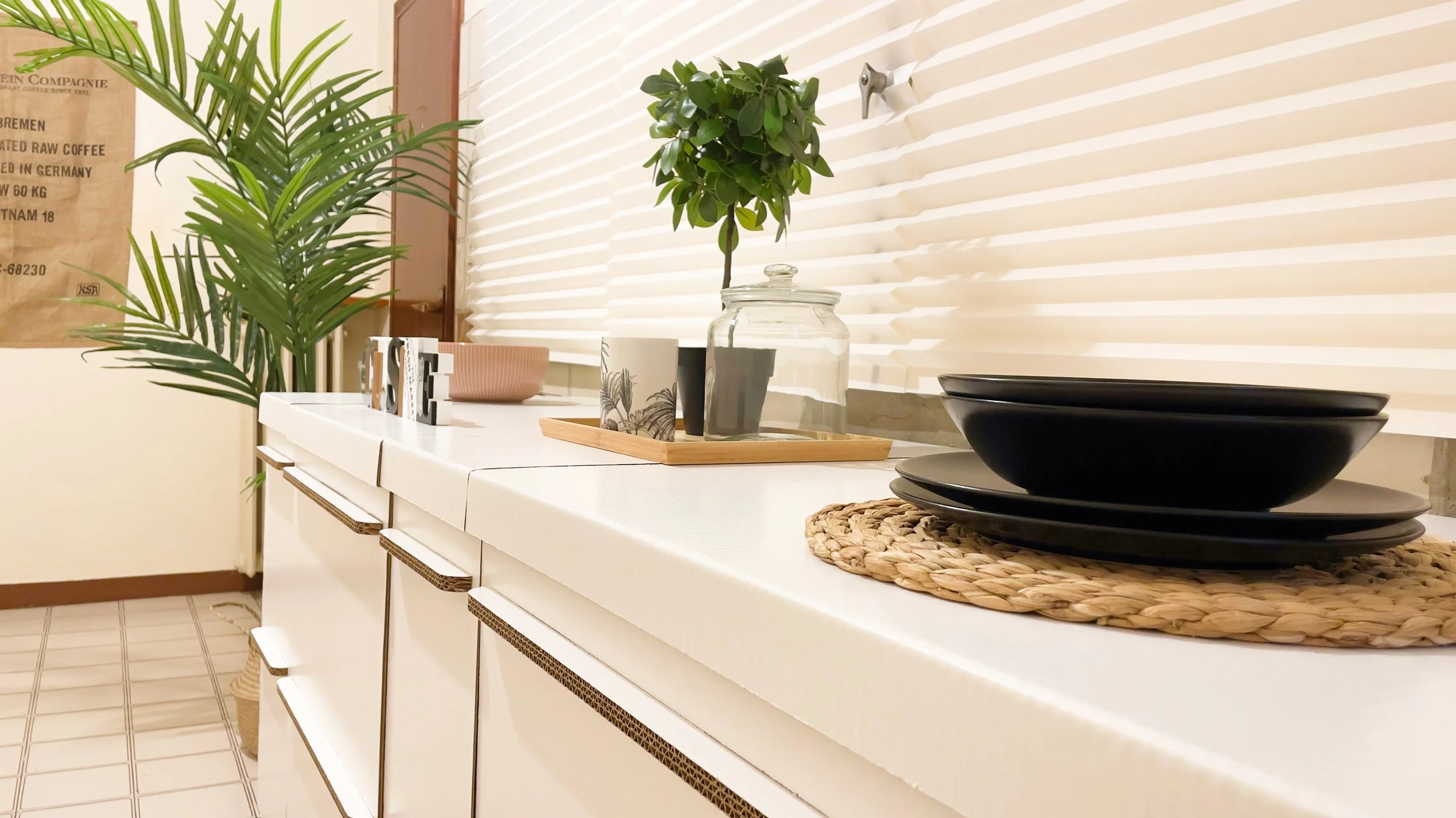 Cucina moderna con piano di lavoro bianco, stoviglie nere su sottopiatto intrecciato, vassoio con tazza e pianta decorativa, pianta alta nell'angolo, parete con tenda a persiana.