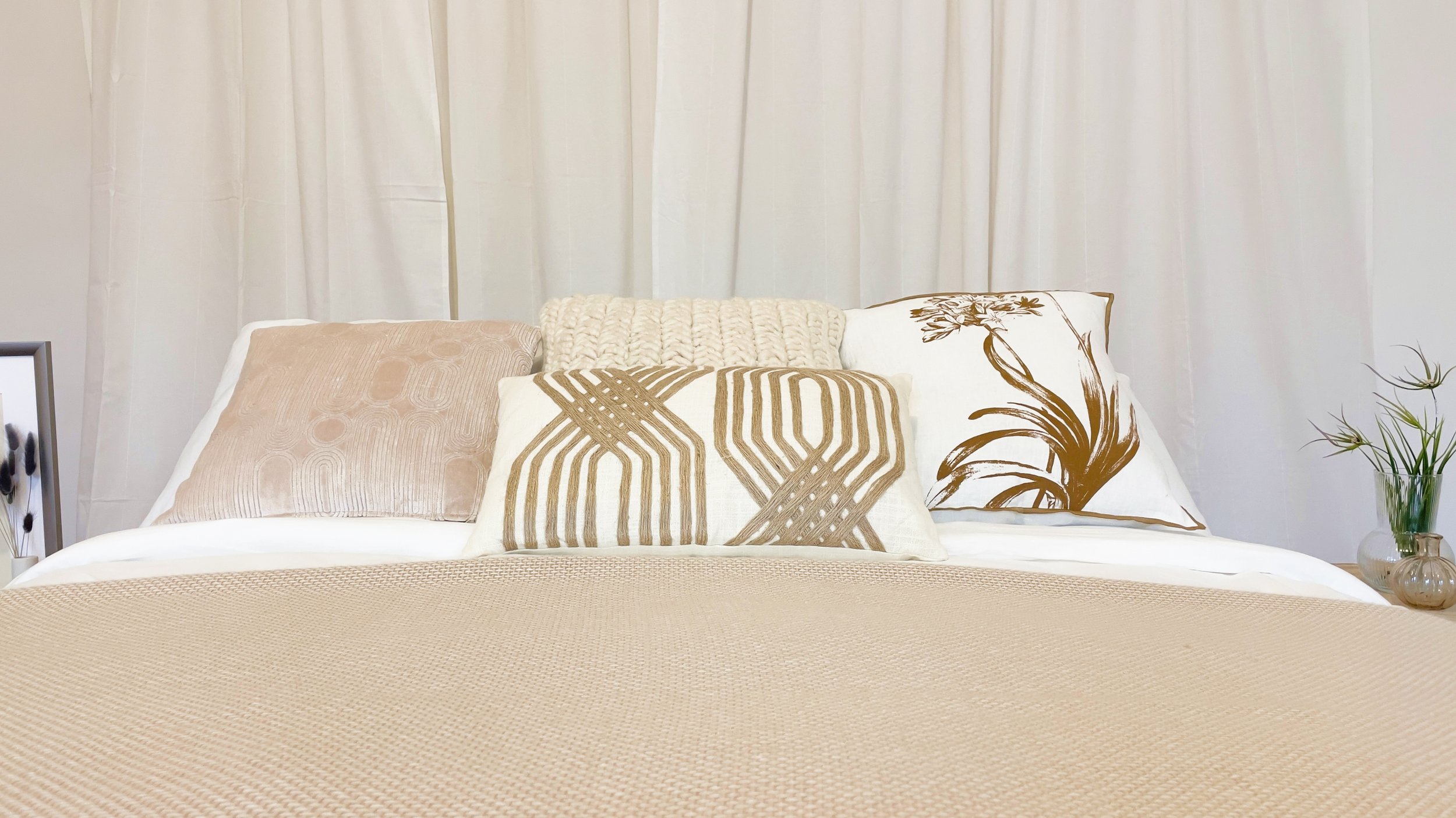 Letto con cuscini decorativi e coperta beige, sfondo con tende bianche e un vaso con pianta a destra.