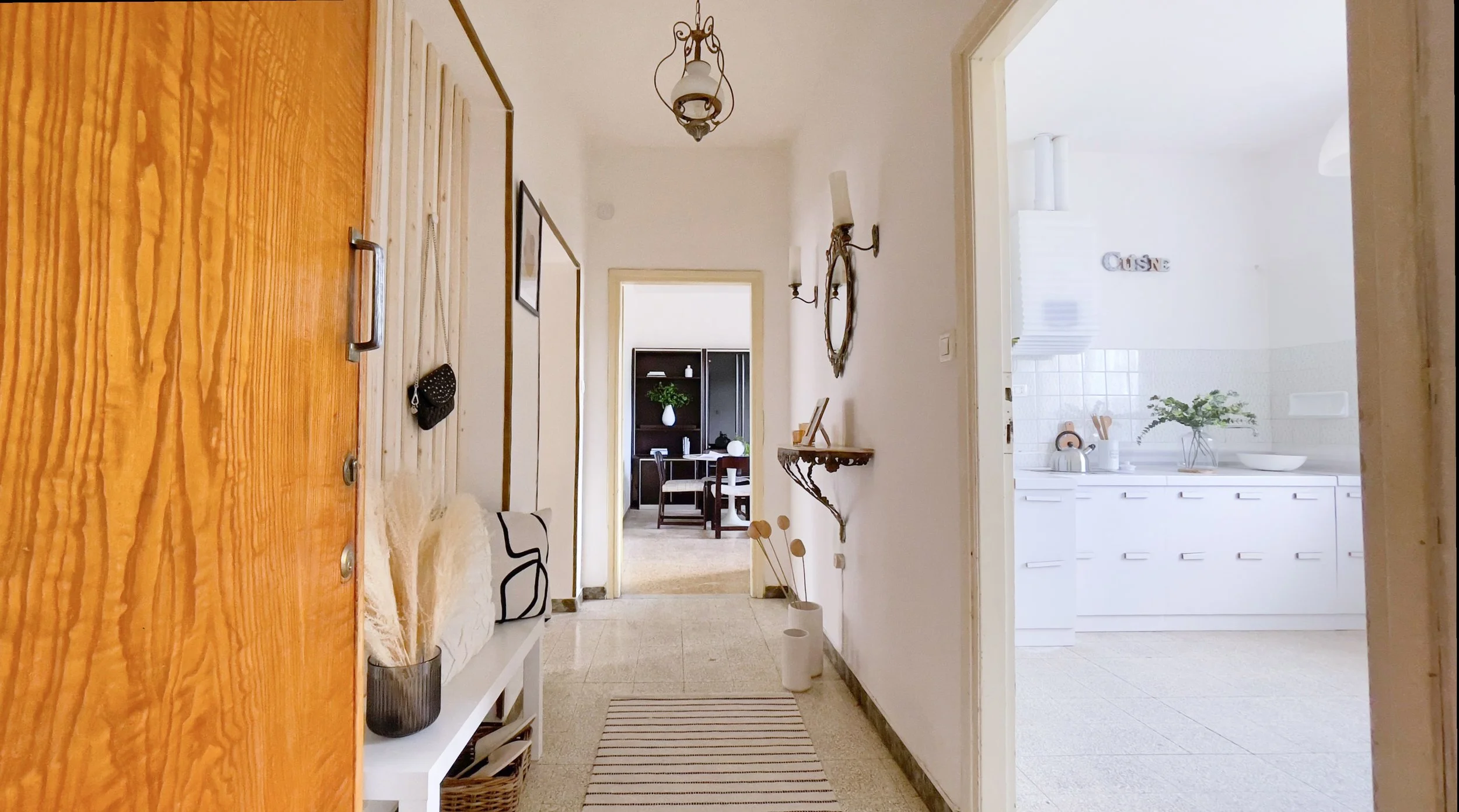 Corridoio luminoso con pareti bianche, pavimento chiaro e tappeto a righe. Sulla destra un mobile con specchio e lampada. A sinistra un armadio in legno e un ripiano con vasi decorativi. In fondo, visibile una cucina bianca e una sala da pranzo con m
