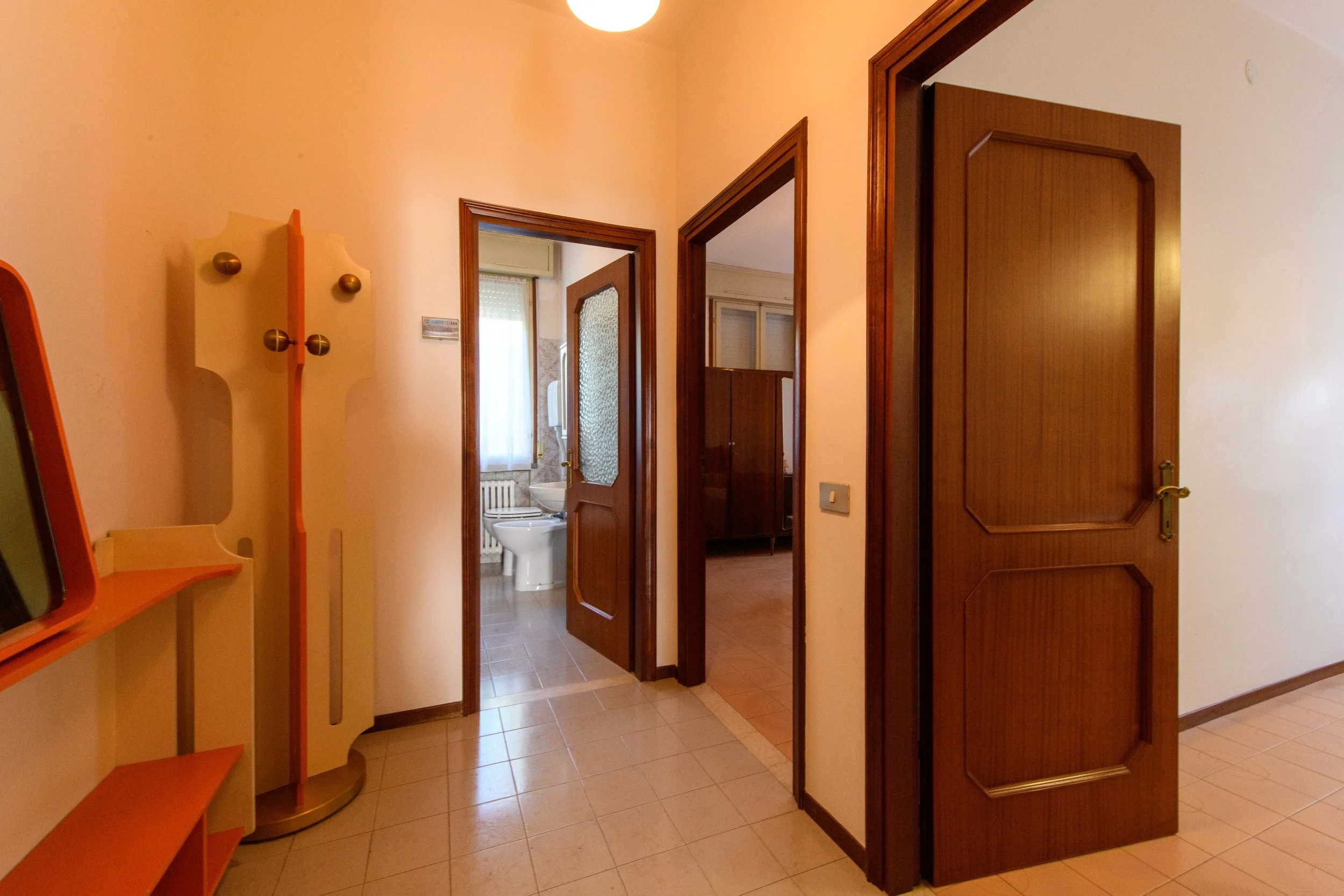 Interno di un appartamento con corridoio, due porte aperte che conducono a stanze diverse, un tavolino e appendiabiti. Vista del bagno sullo sfondo.