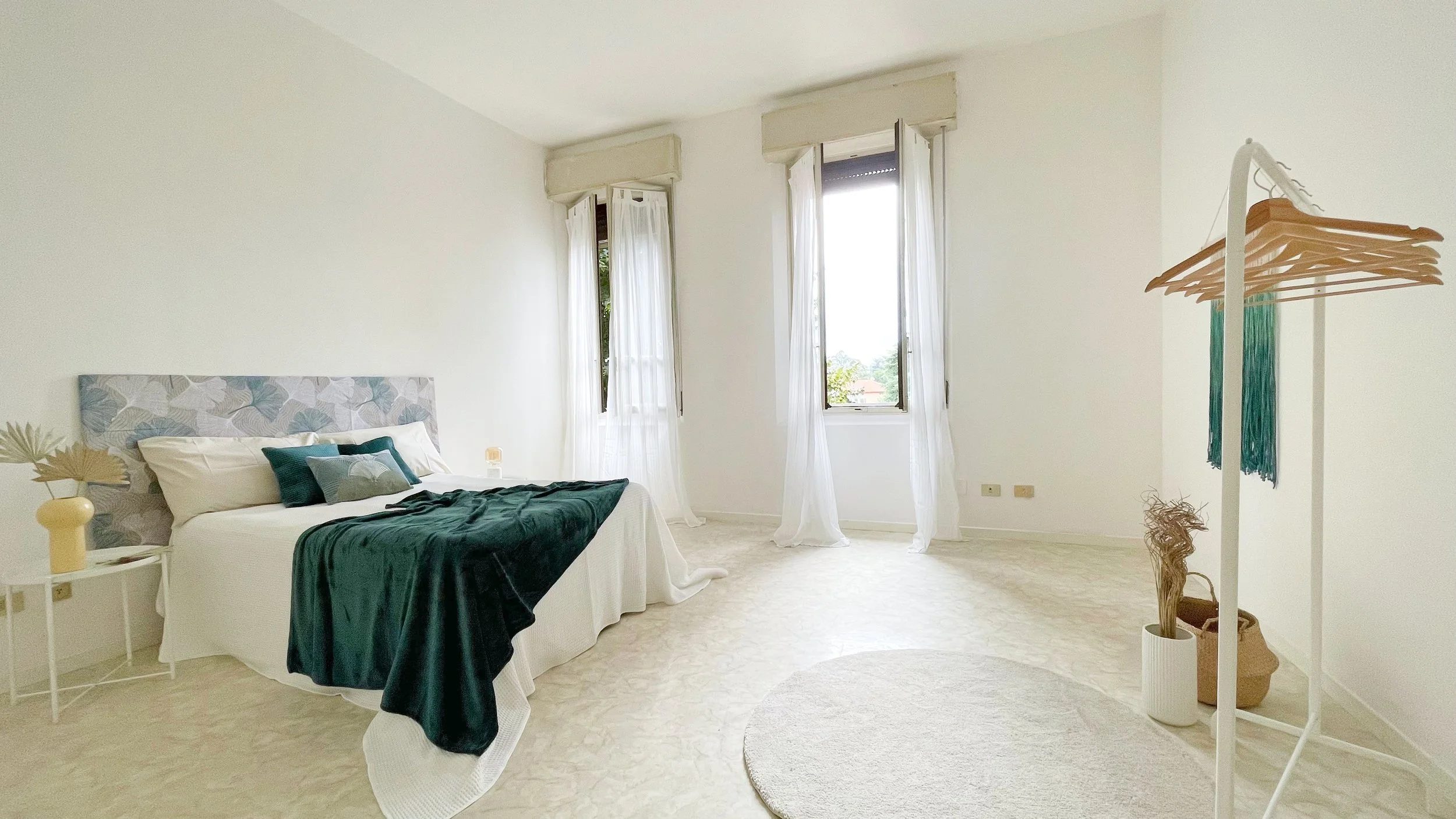 Camera da letto minimalista con letto matrimoniale, coperte verdi, finestre luminose, tenda in legno con stampelle e decorazioni minimaliste.