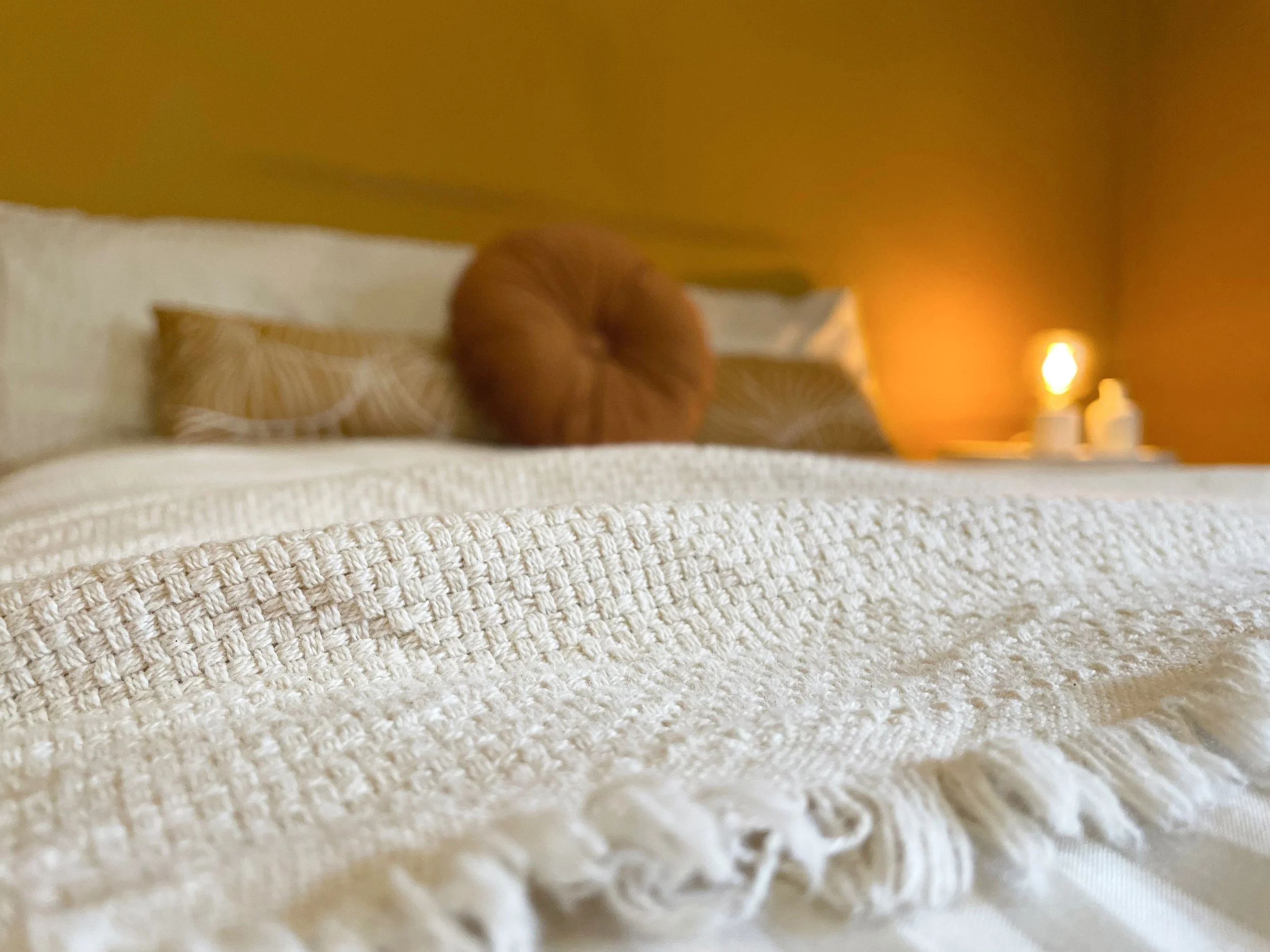Letto con coperta bianca e cuscini decorativi, illuminato da una lampada su un comodino.