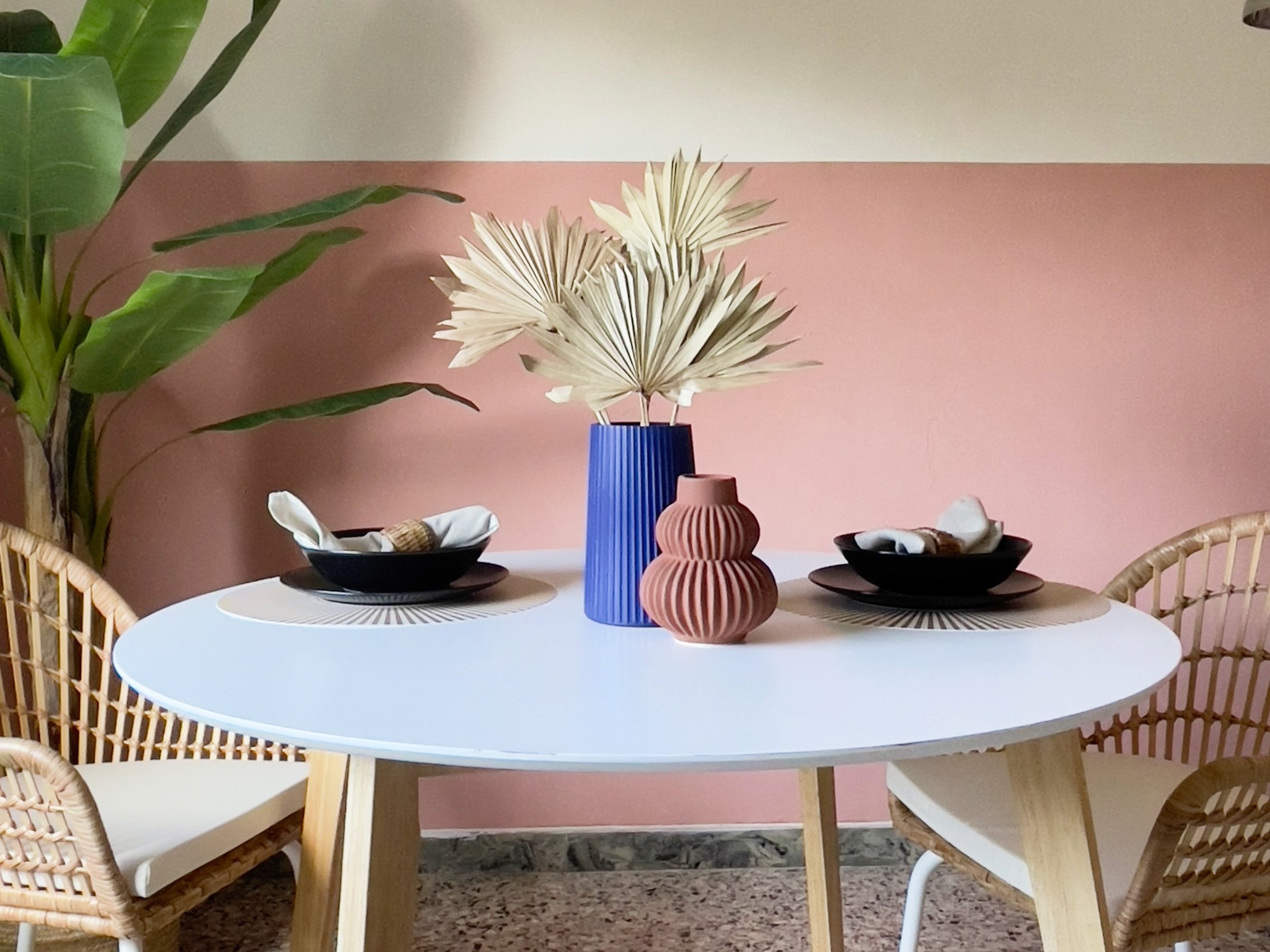 Tavolo rotondo bianco con decorazioni, due sedie intrecciate, piatti neri con tovaglioli, vasi blu e rosso, foglie decorative, sfondo rosa e bianco.