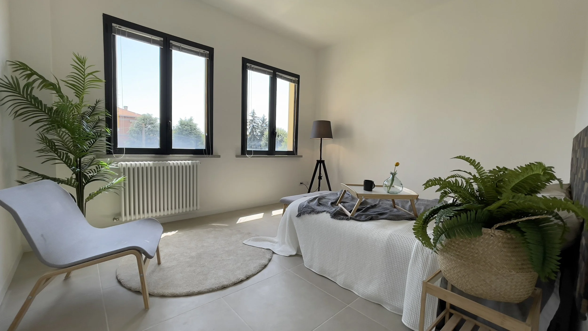 Camera minimalista con grandi finestre, una sedia moderna, una lampada da terra, piante verdi e un letto semplice con un vassoio. Design luminoso e accogliente.