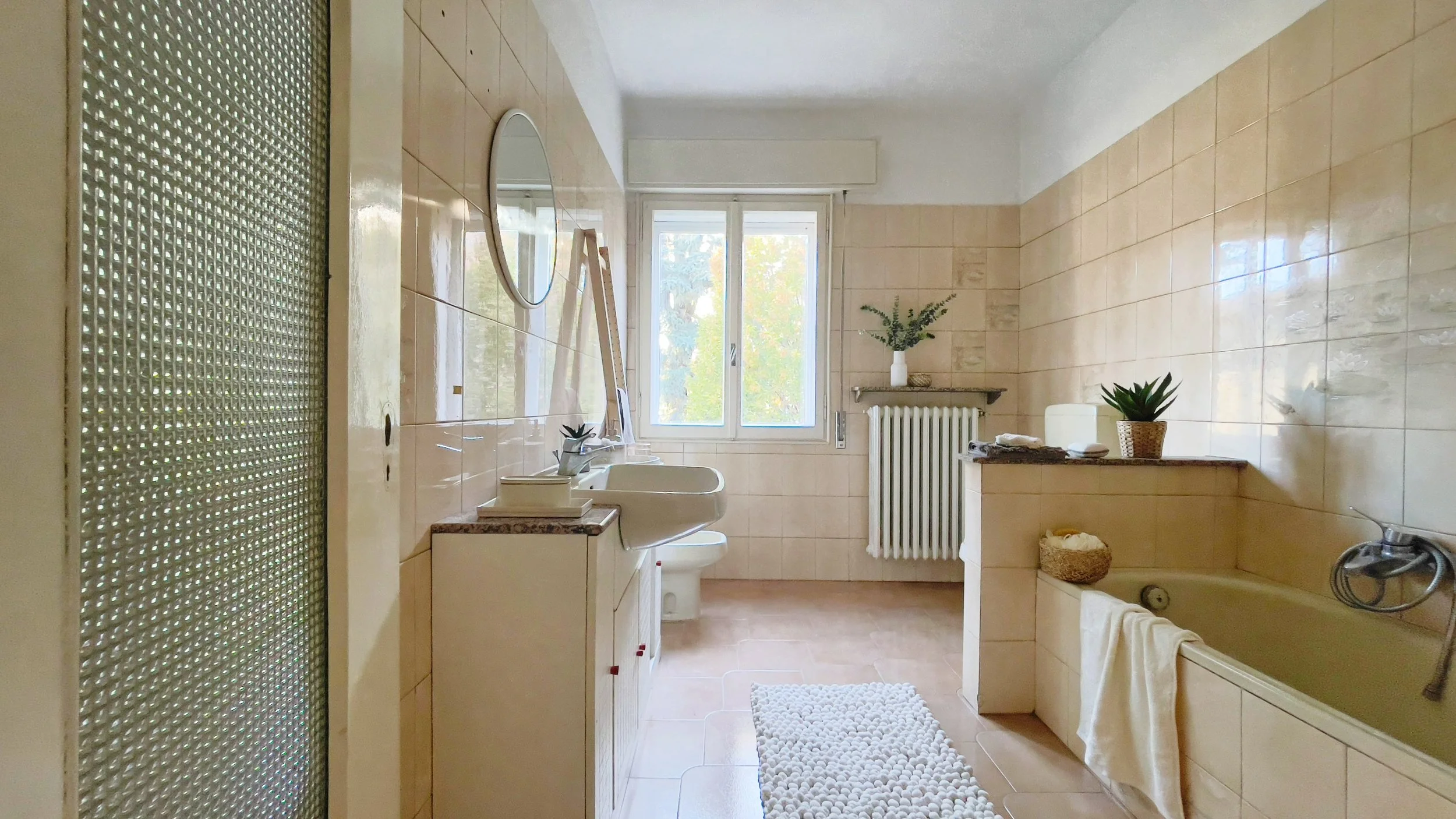 Bagno moderno con piastrelle beige, specchio rotondo, finestra con luce naturale, lavandino, vasca da bagno e piante decorative.