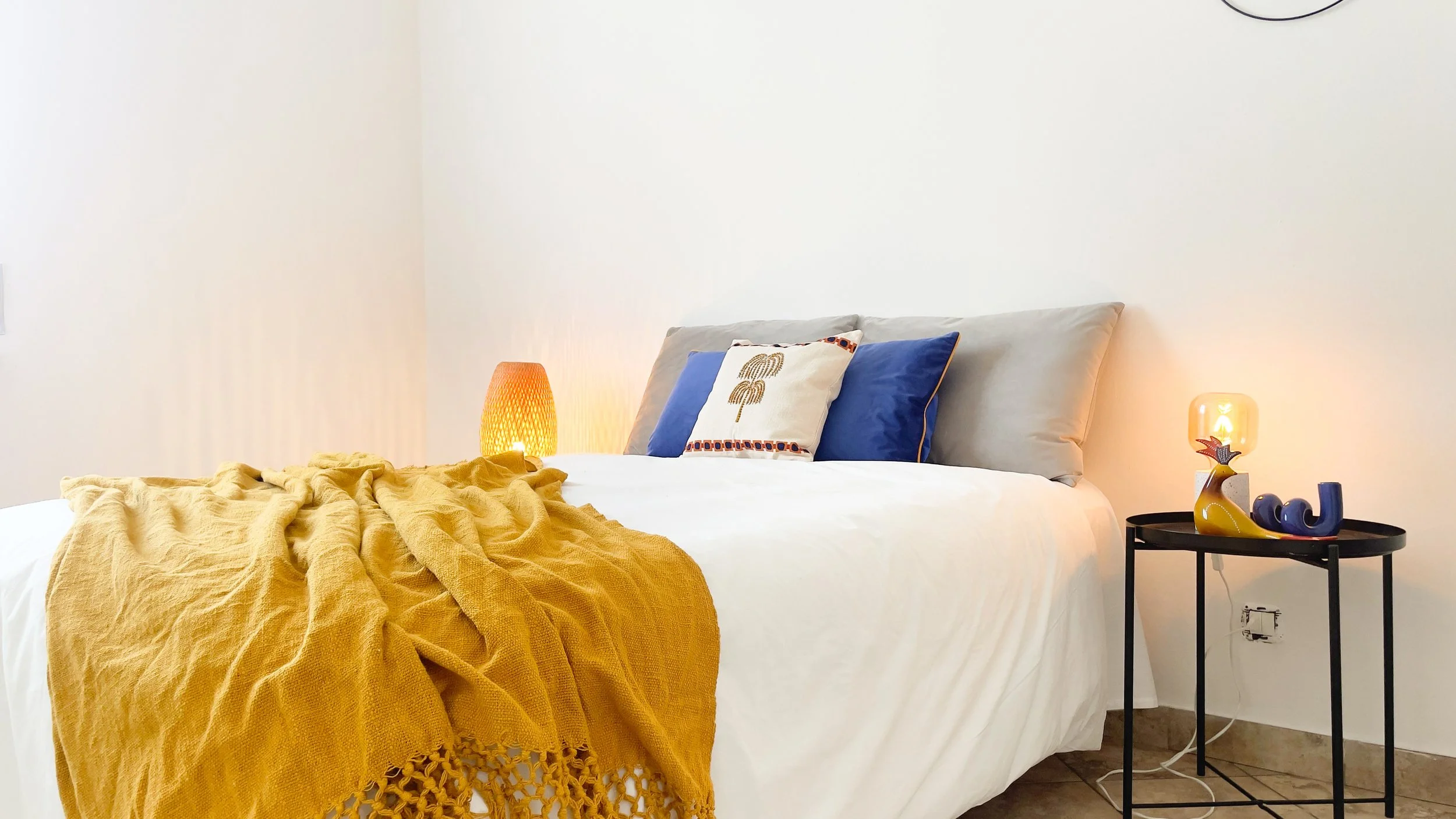 Camera da letto moderna con letto bianco, coperta gialla, cuscini decorativi, lampade sul comodino e pareti bianche.