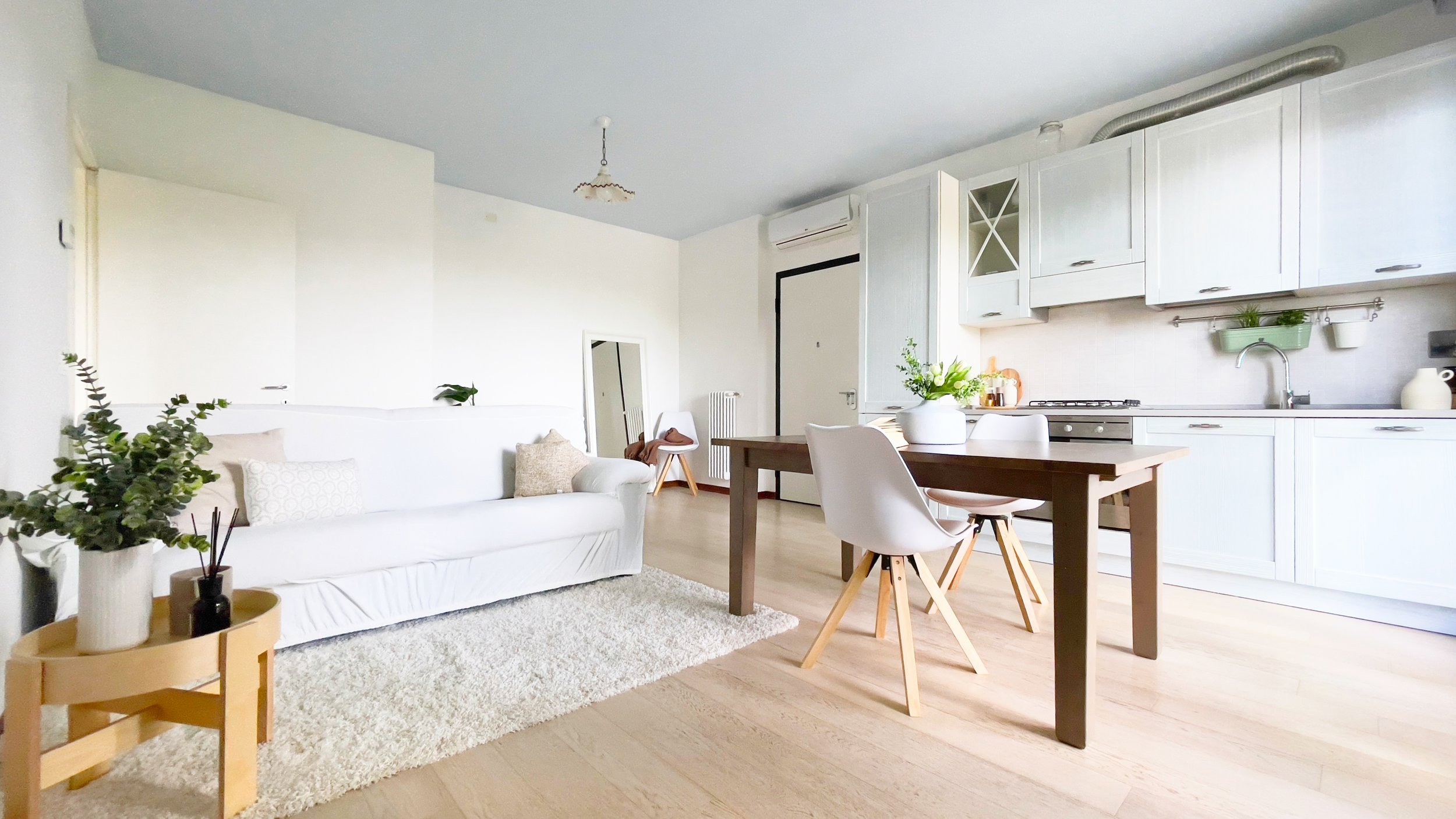 Soggiorno moderno con cucina a vista, divano bianco, tavolo da pranzo in legno scuro e sedie, pareti bianche e pavimento in legno chiaro, elementi decorativi e piante.