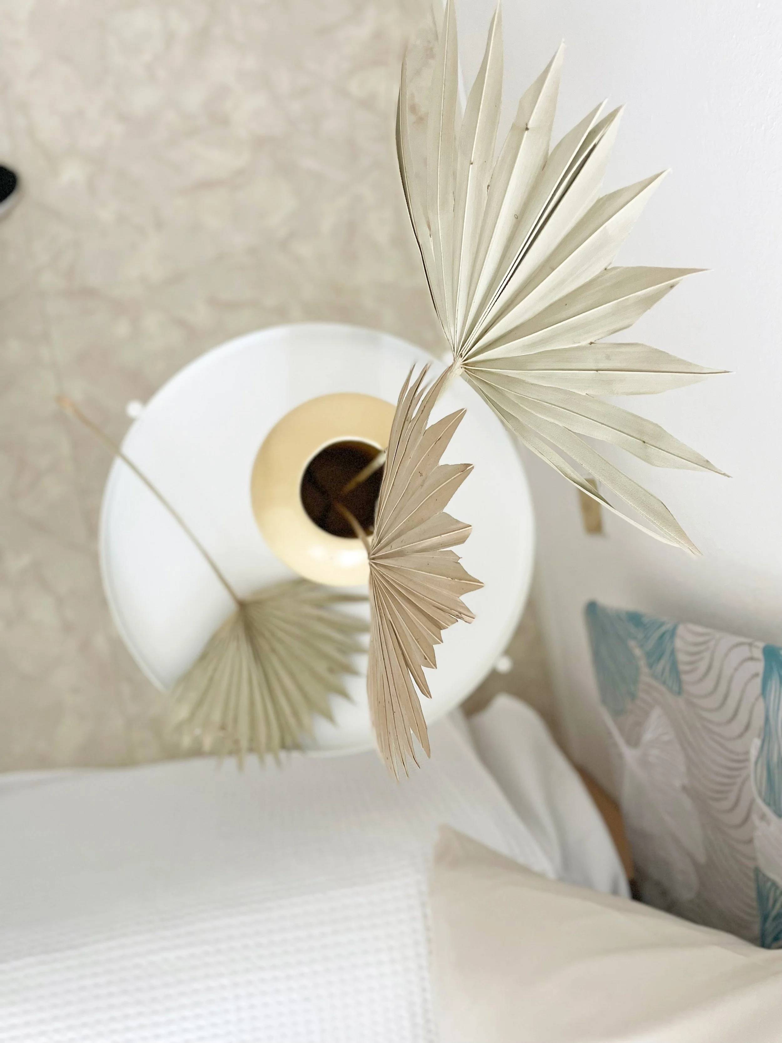 Vista dall'alto di un tavolo rotondo con un vaso che contiene foglie di palma essiccate, accanto a un letto con cuscini colorati.