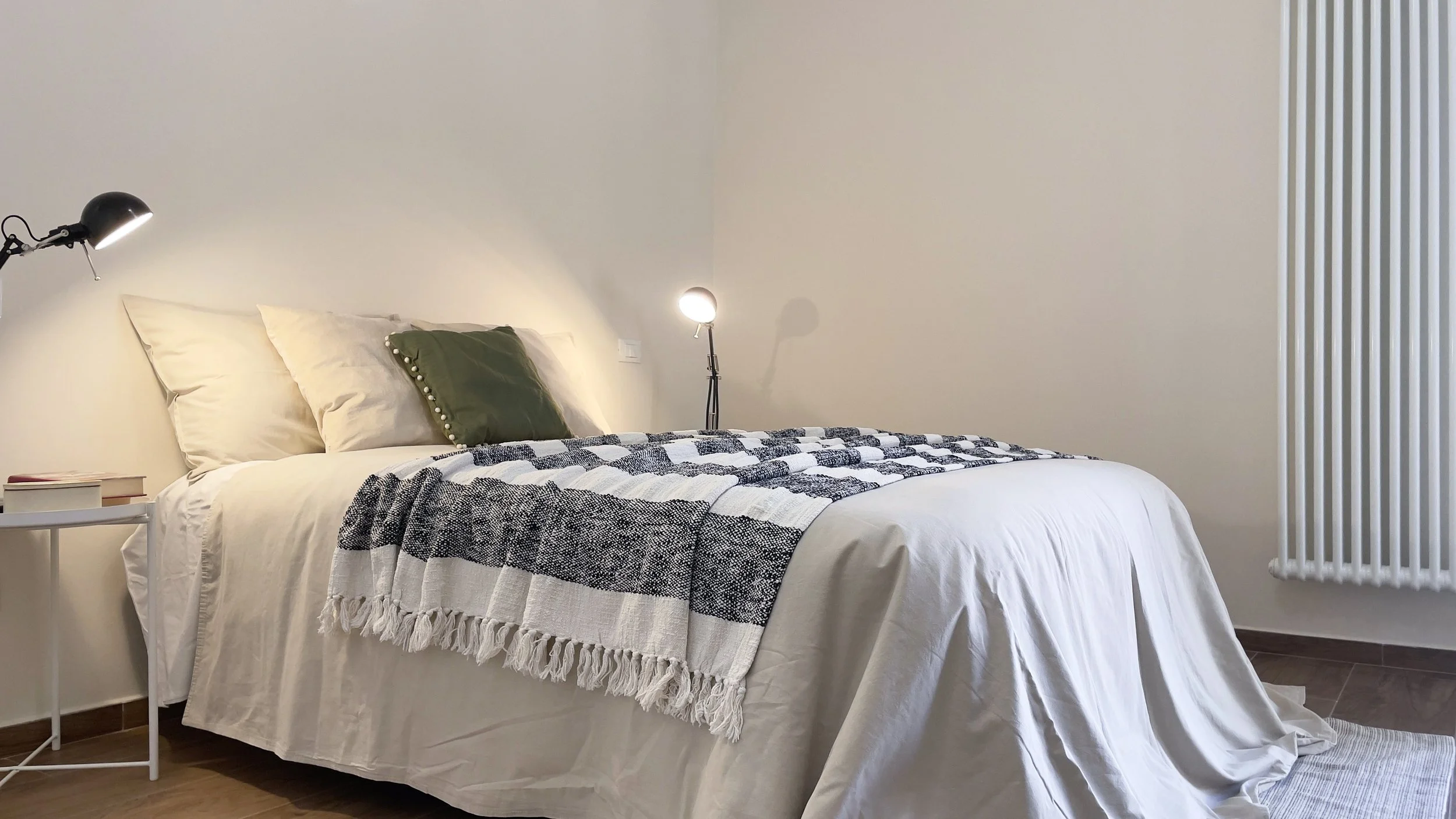Camera da letto moderna con letto matrimoniale, coperta a scacchi bianchi e neri, cuscini, lampada da tavolo nera su un comodino bianco, pavimento in legno, parete beige, e calorifero bianco verticale.