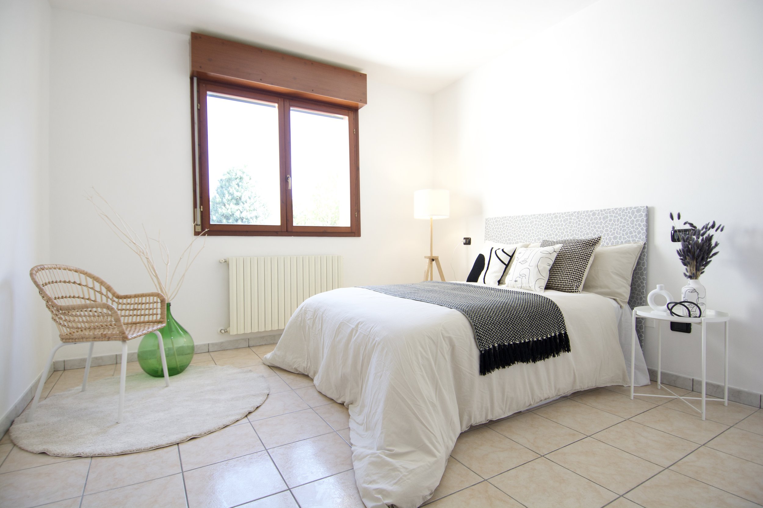 Camera da letto moderna con letto matrimoniale, pareti bianche, moquette chiara, sedia in rattan, lampada da terra, comodino con decorazioni e finestra luminosa.
