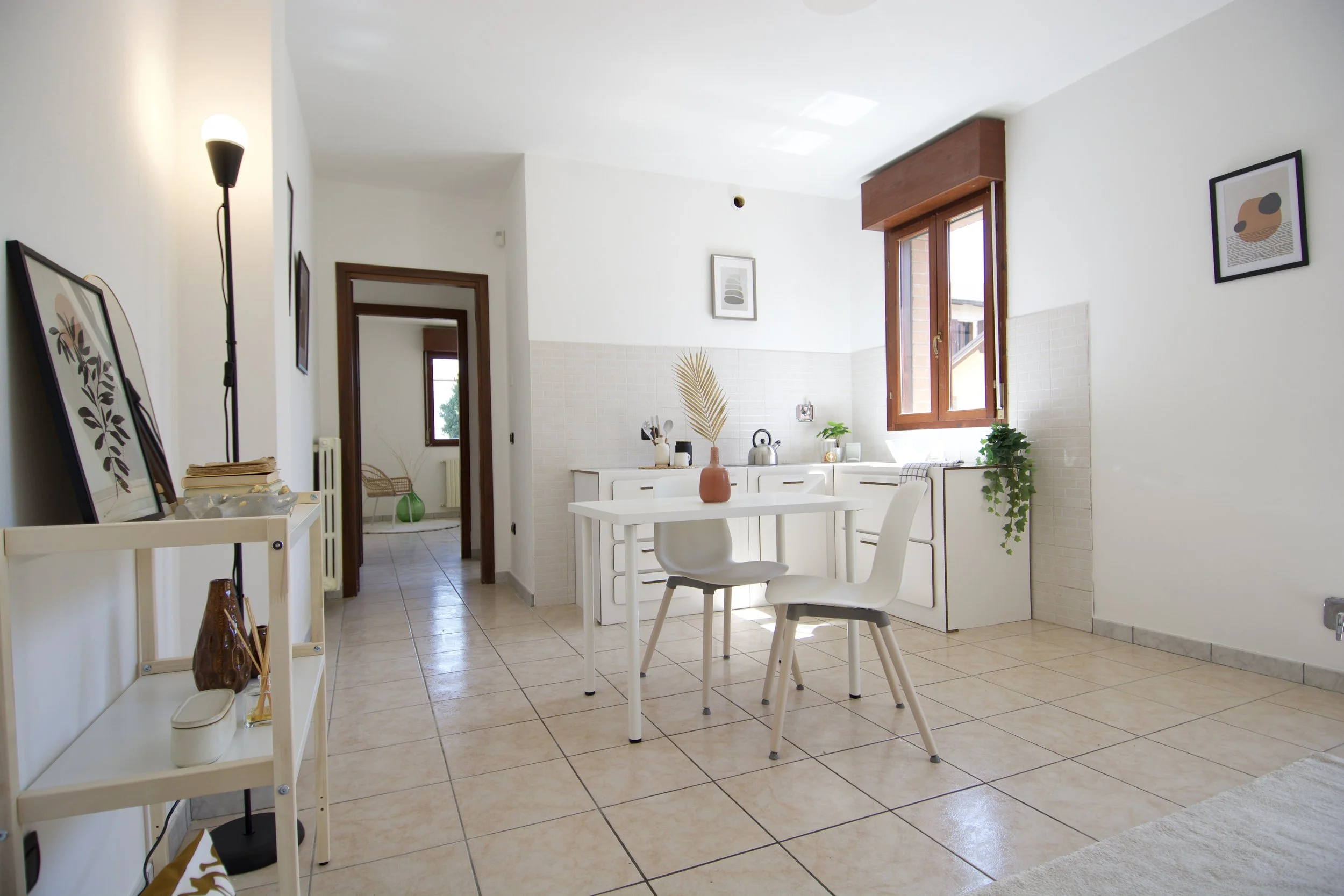 Interno di una cucina moderna e minimalista con pareti bianche. Al centro c'è un tavolo bianco con due sedie. Sulla sinistra, una mensola con oggetti decorativi. A destra, una finestra luminosa con piante. Si intravede un altro ambiente attraverso un