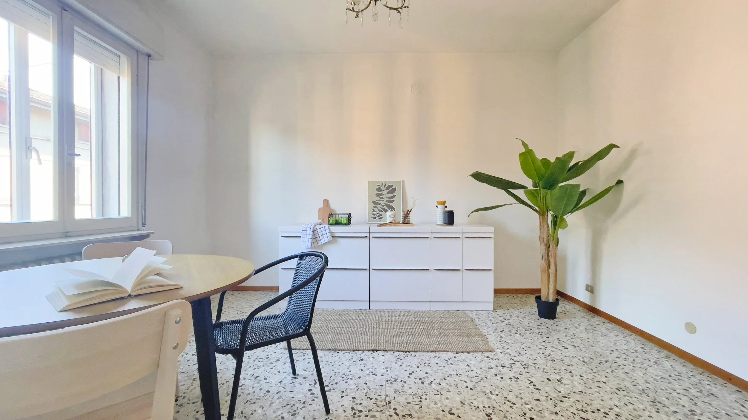 Interno luminoso di una stanza con un tavolo rotondo e due sedie, una credenza bianca con decorazioni, una pianta in vaso, e una finestra grande.