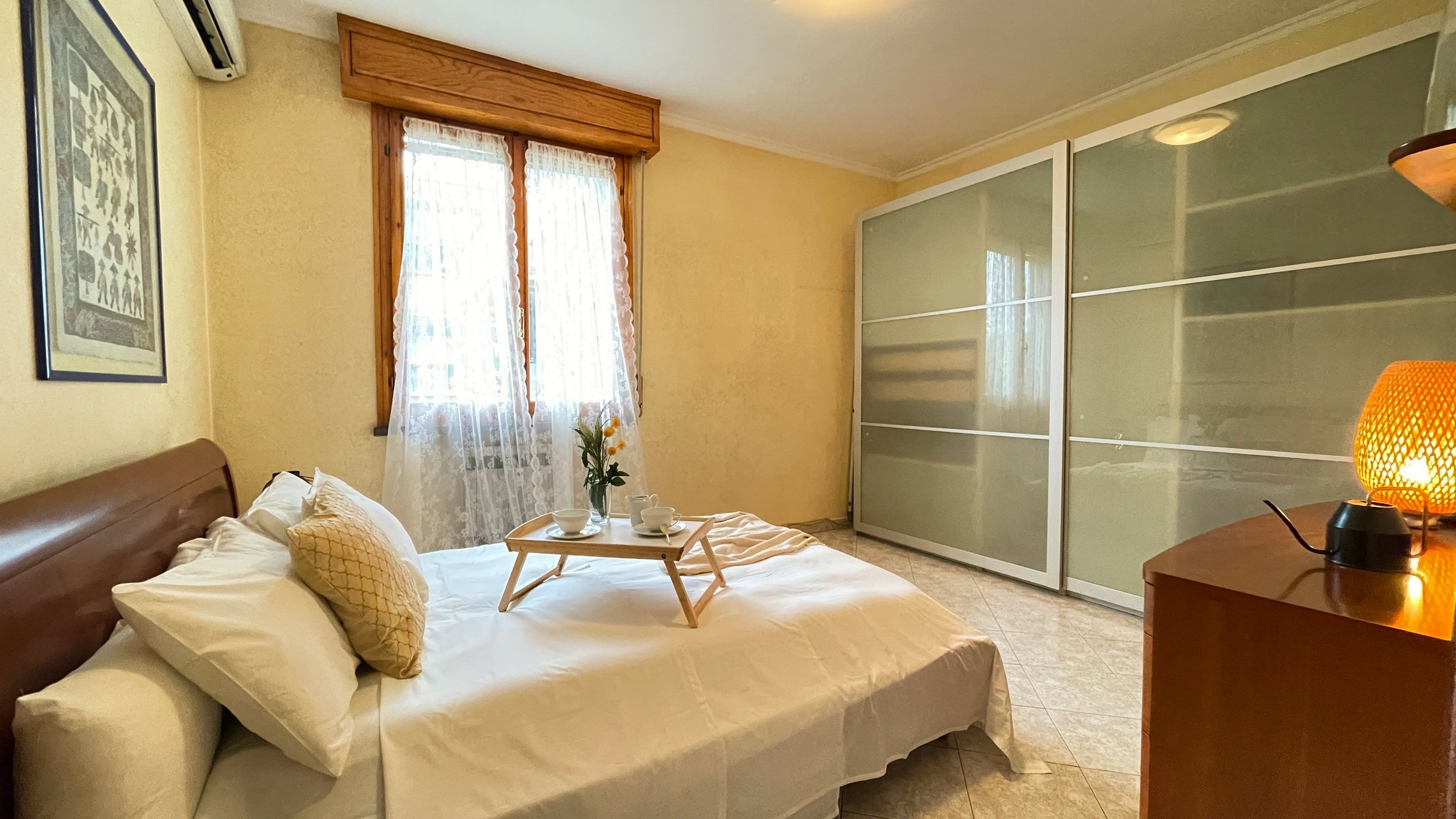 Camera da letto con letto matrimoniale, pareti chiare, armadio con ante scorrevoli in vetro, finestra con tende, quadro appeso e comodino con lampada."}