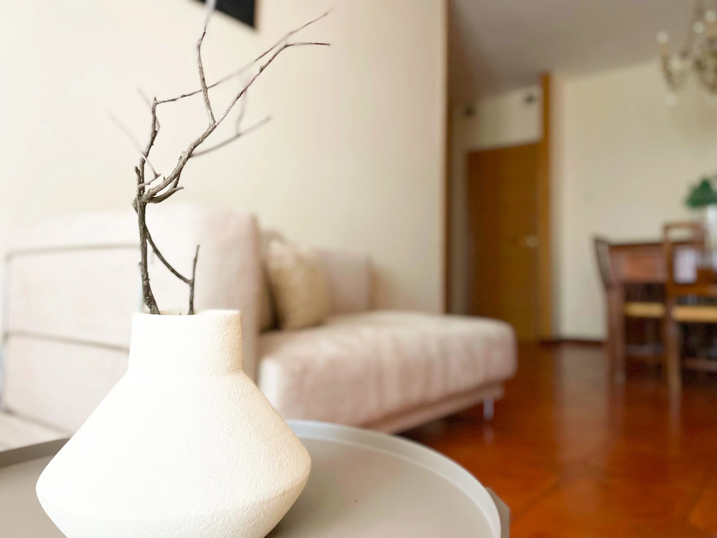 Interno di un soggiorno moderno con divano beige e tavolo in legno, vaso bianco con rami secchi in primo piano.