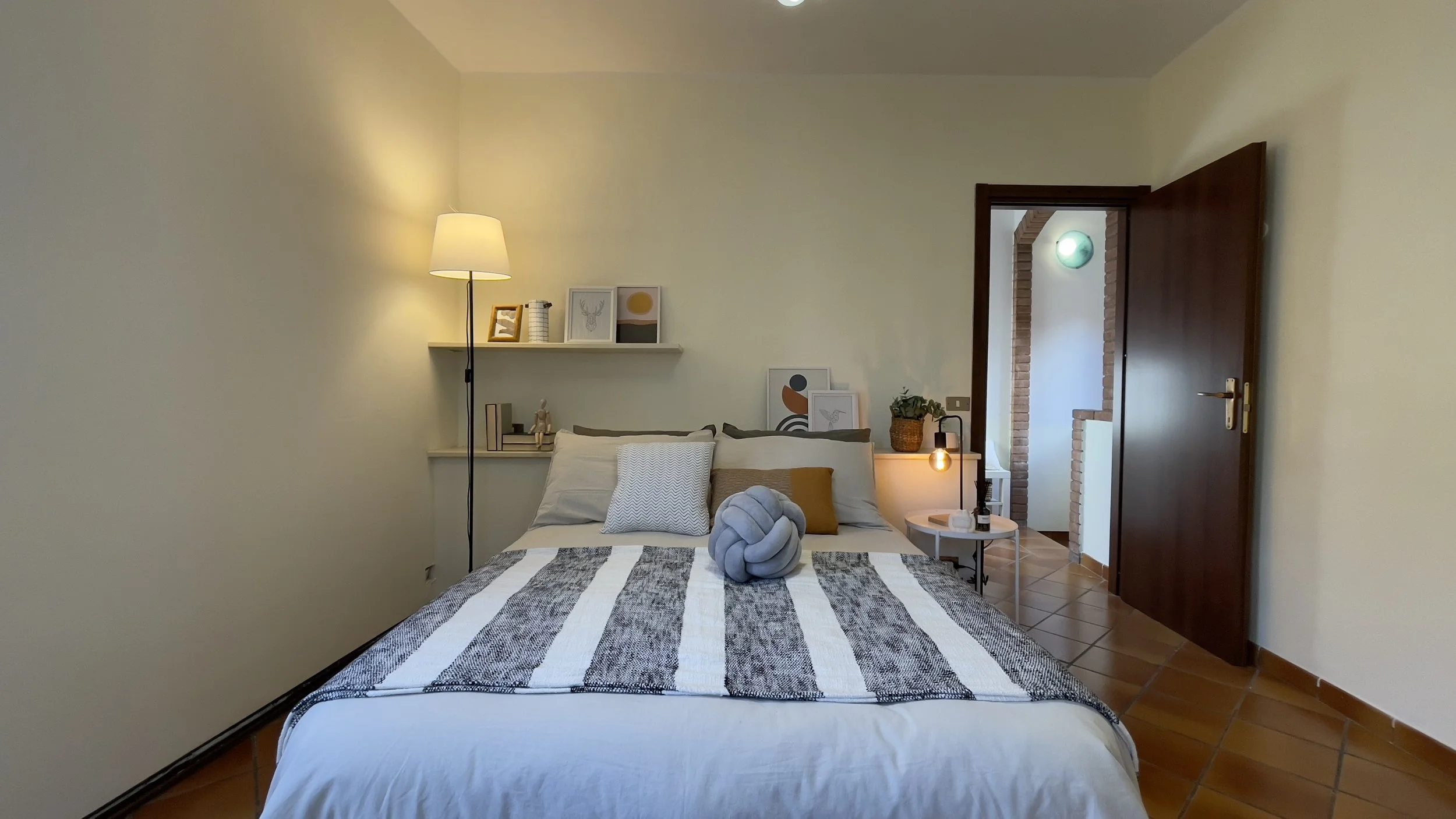Camera da letto con letto matrimoniale, lampada da terra, mensole con decorazioni, parete chiara e pavimento in piastrelle, porta aperta che mostra un corridoio in mattoni.