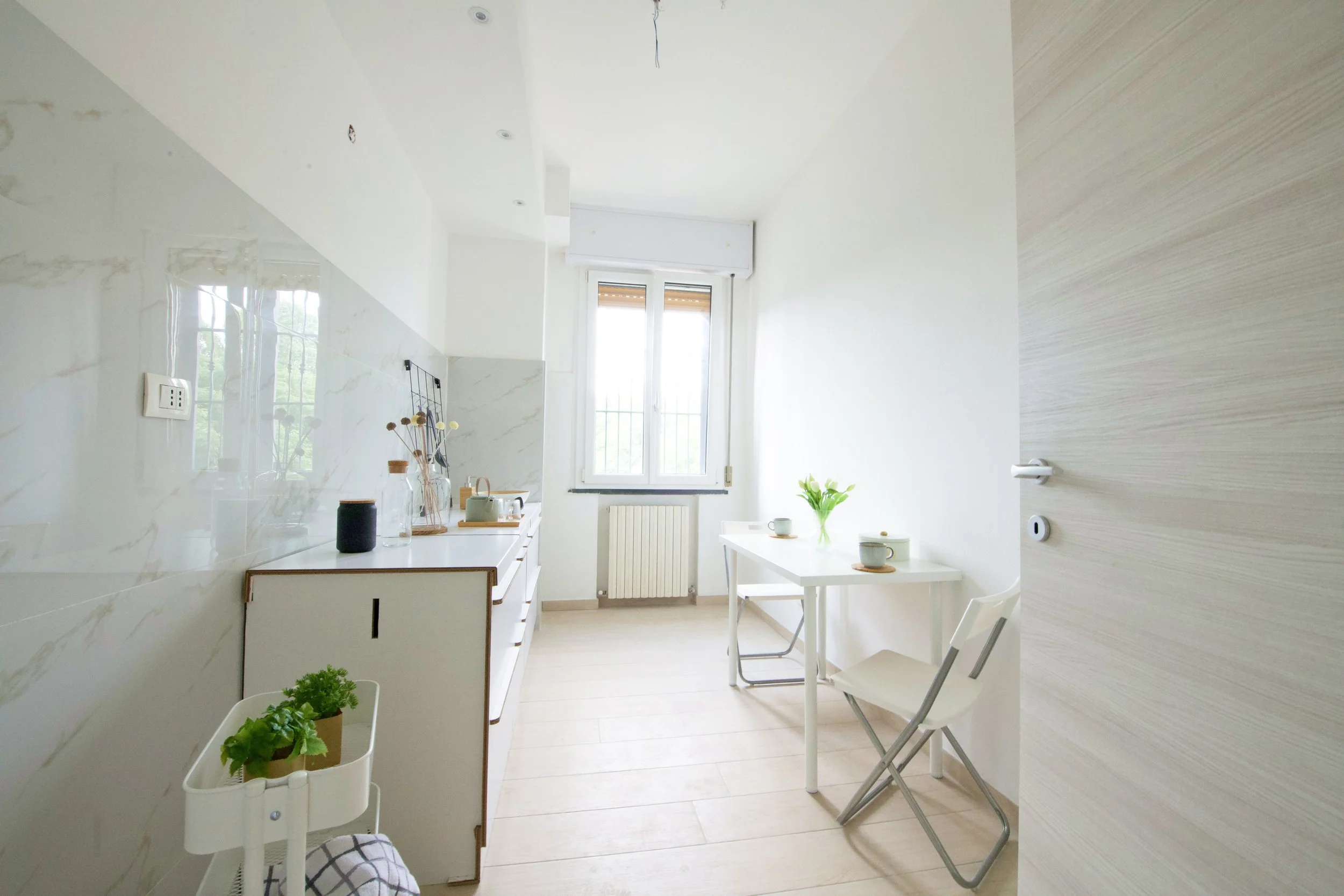 Cucina moderna e luminosa con mobili bianchi, un tavolo con sedie pieghevoli, piante e stoviglie, e una finestra che lascia entrare luce naturale.