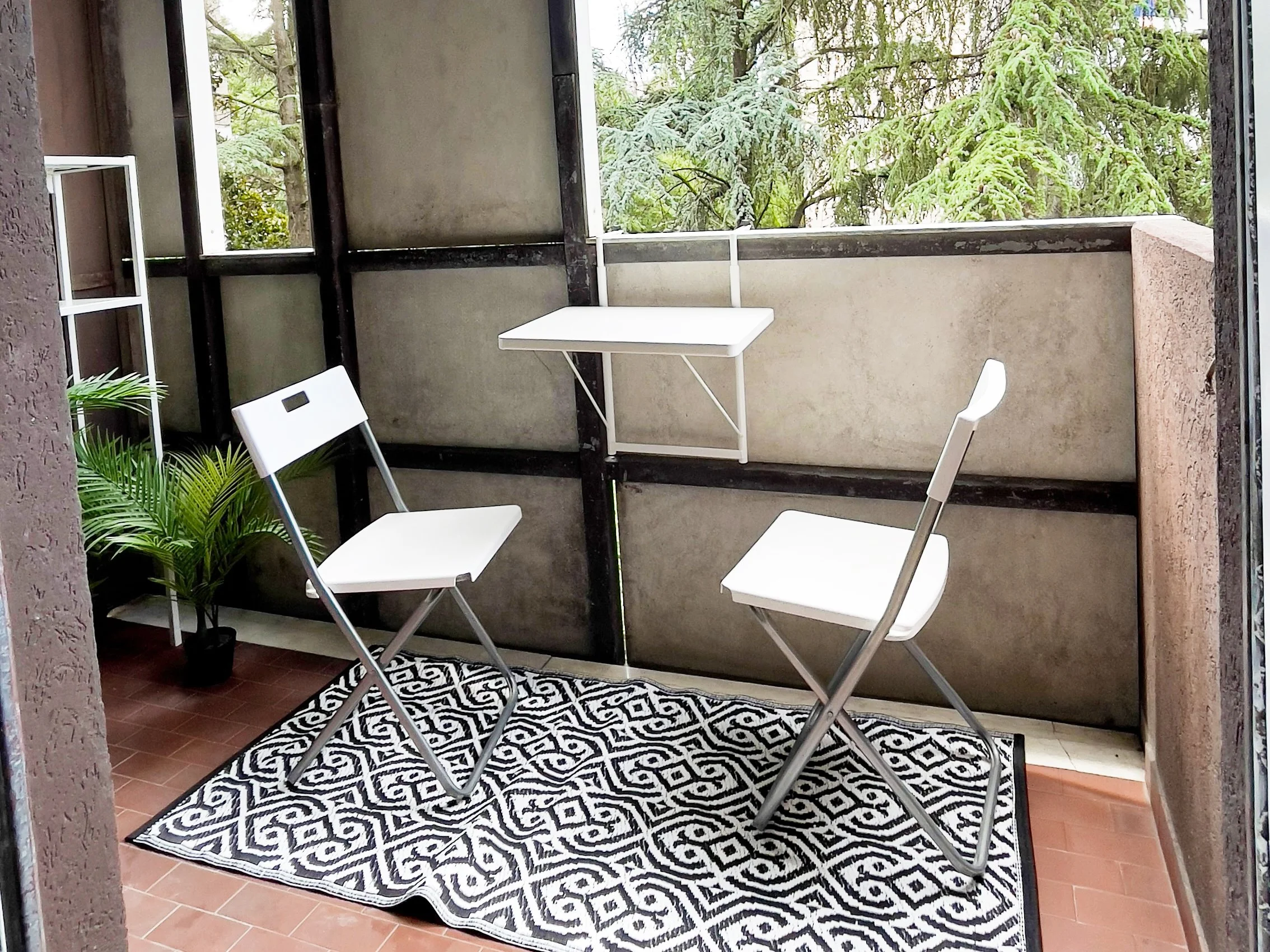 Balcone con due sedie pieghevoli bianche, un tavolino sospeso, tappeto in bianco e nero con motivi geometrici, pianta in vaso e scaffale bianco.