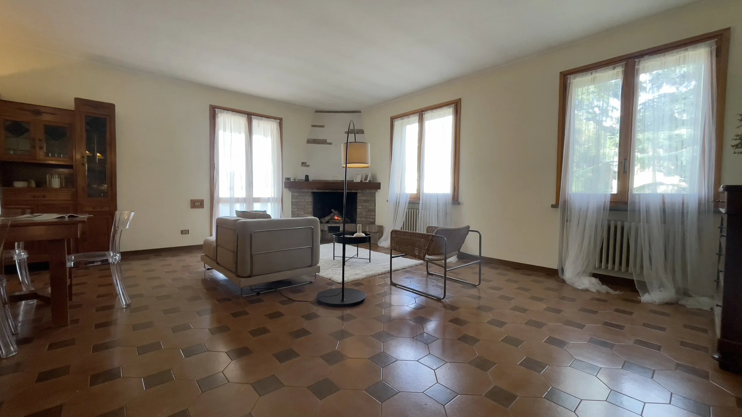 Salotto con pavimento in piastrelle marroni, camino, due poltrone, tavolino, e finestra con tende bianche trasparenti.