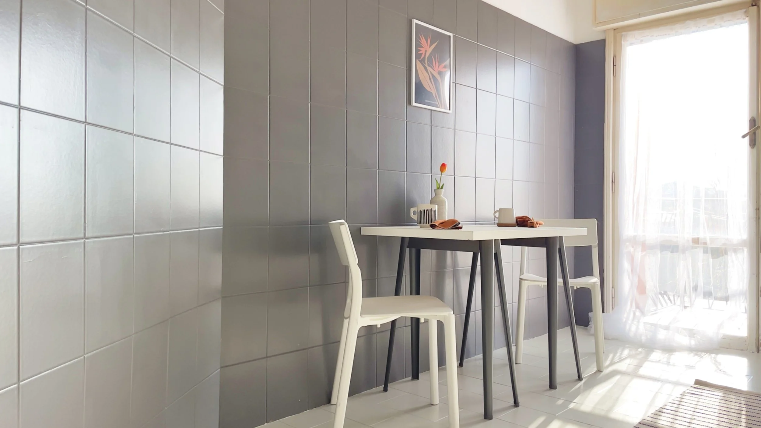 Interno di una cucina moderna con una parete piastrellata grigia, un tavolo bianco e due sedie bianche. Sul tavolo ci sono una tazza, un vaso con un fiore e dei tovaglioli. La luce solare entra da una finestra con tende bianche.