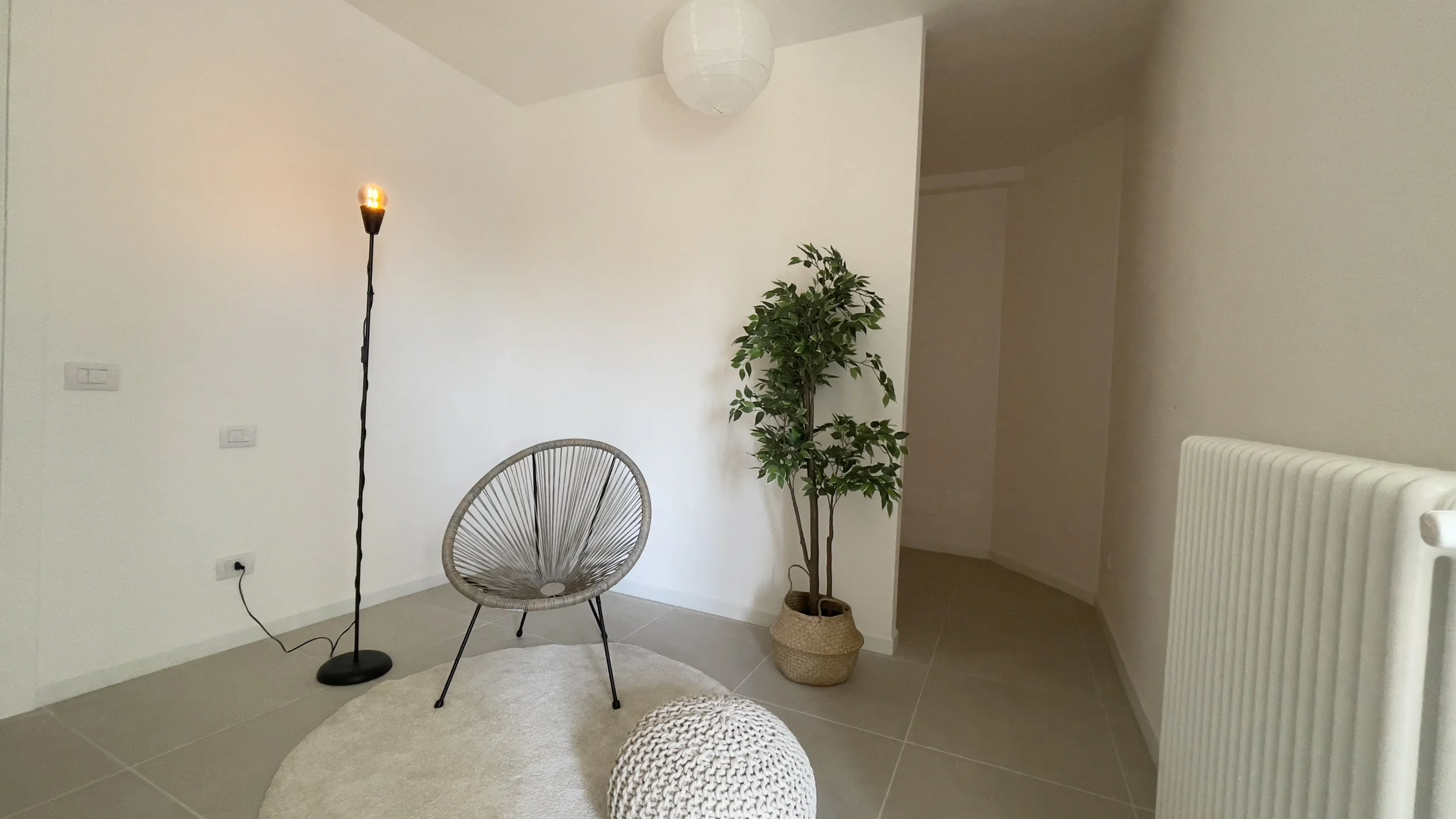 Interno di una stanza minimalista con una sedia moderna, lampada da terra, tappeto rotondo, e pianta in vaso. Pareti bianche e pavimento piastrellato.