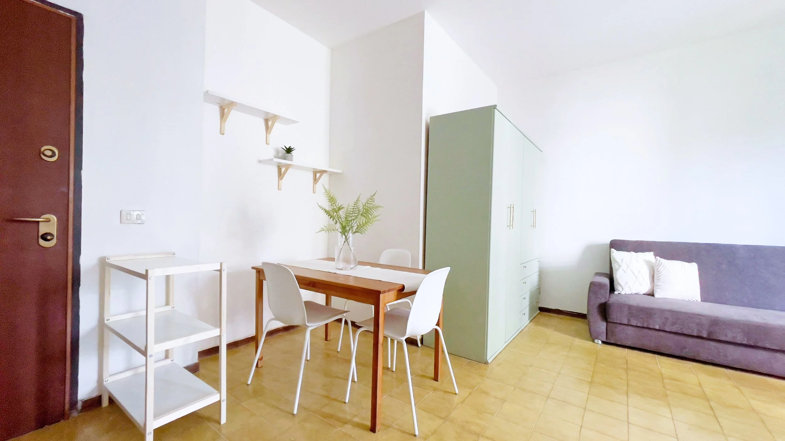 Interno di una stanza luminosa con tavolo in legno e sedie bianche. C'è un divano grigio con cuscini bianchi, un armadio verde chiaro, e mensole con decorazioni. A sinistra, una porta d'ingresso marrone chiaro con una piccola scaffalatura bianca. Su 