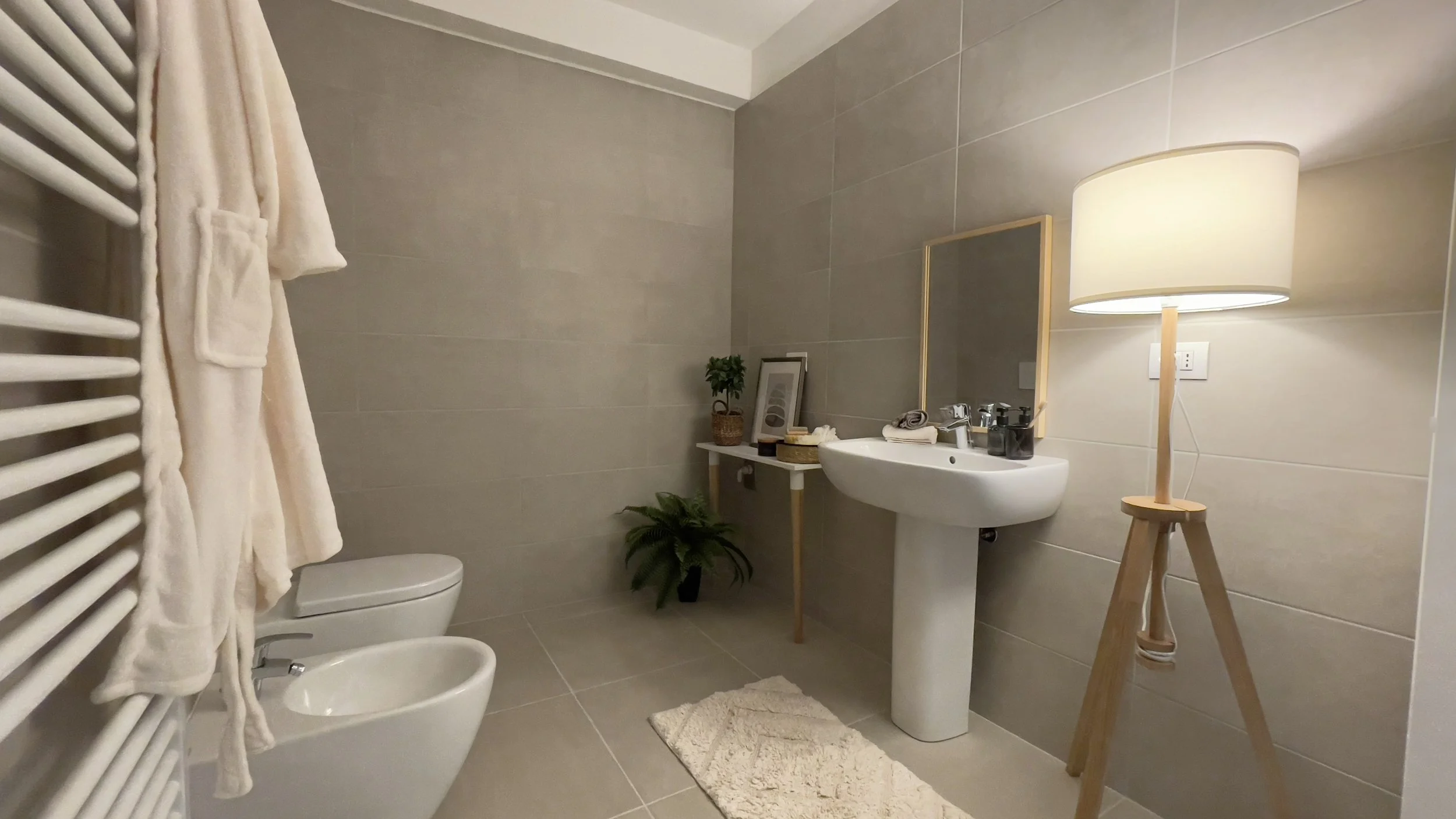 Bagno moderno con piastrelle grigie, lavabo, specchio, bidet e WC. Accappatoio bianco appeso a un termosifone. Tavolino con piante e decorazioni. Lampada da terra.