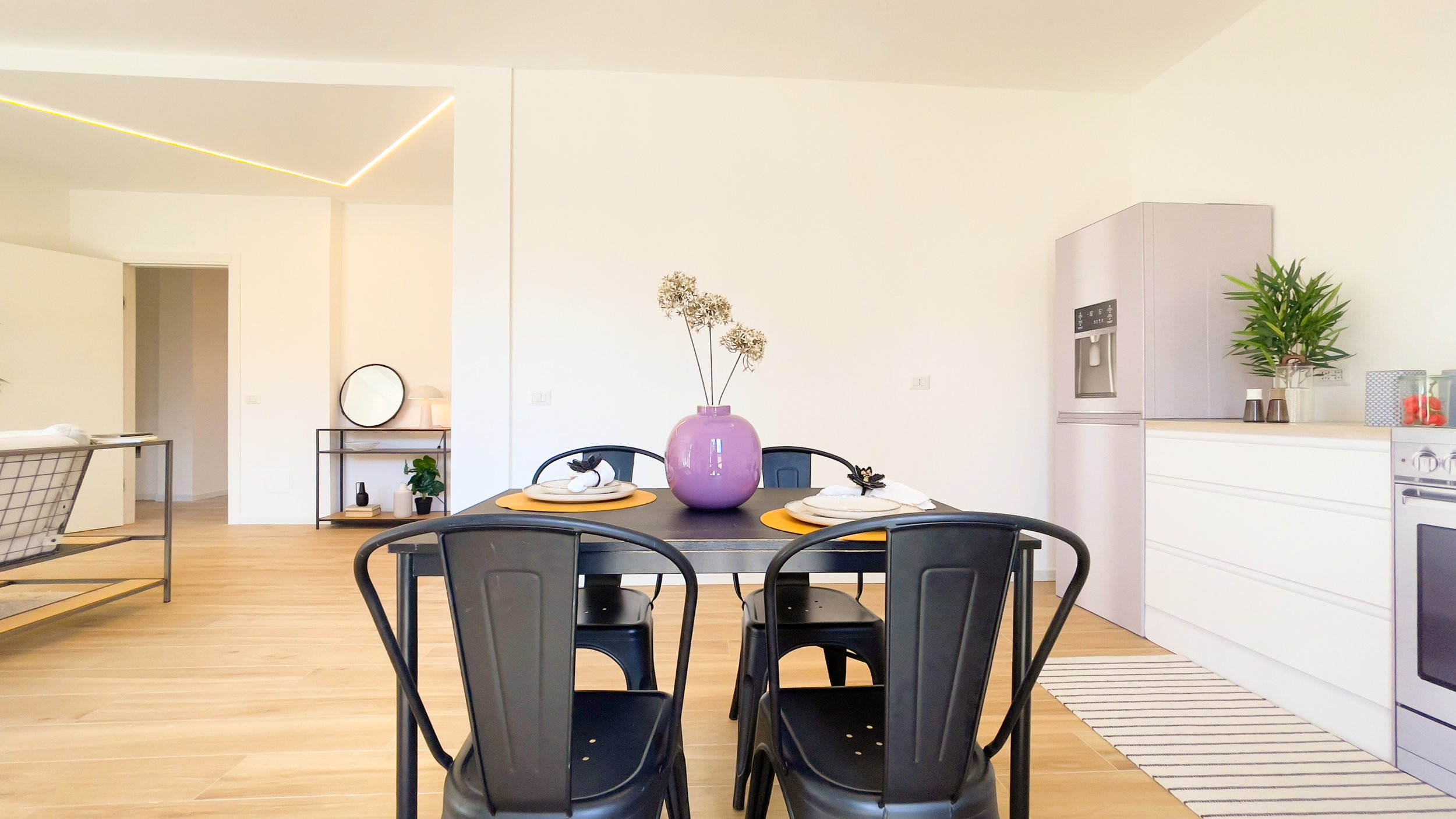 Interno di una cucina moderna con un tavolo da pranzo nero, un vaso viola con fiori secchi, sedie nere, frigorifero grigio e mobili bianchi. Pavimento in legno chiaro e decorazioni minimali.