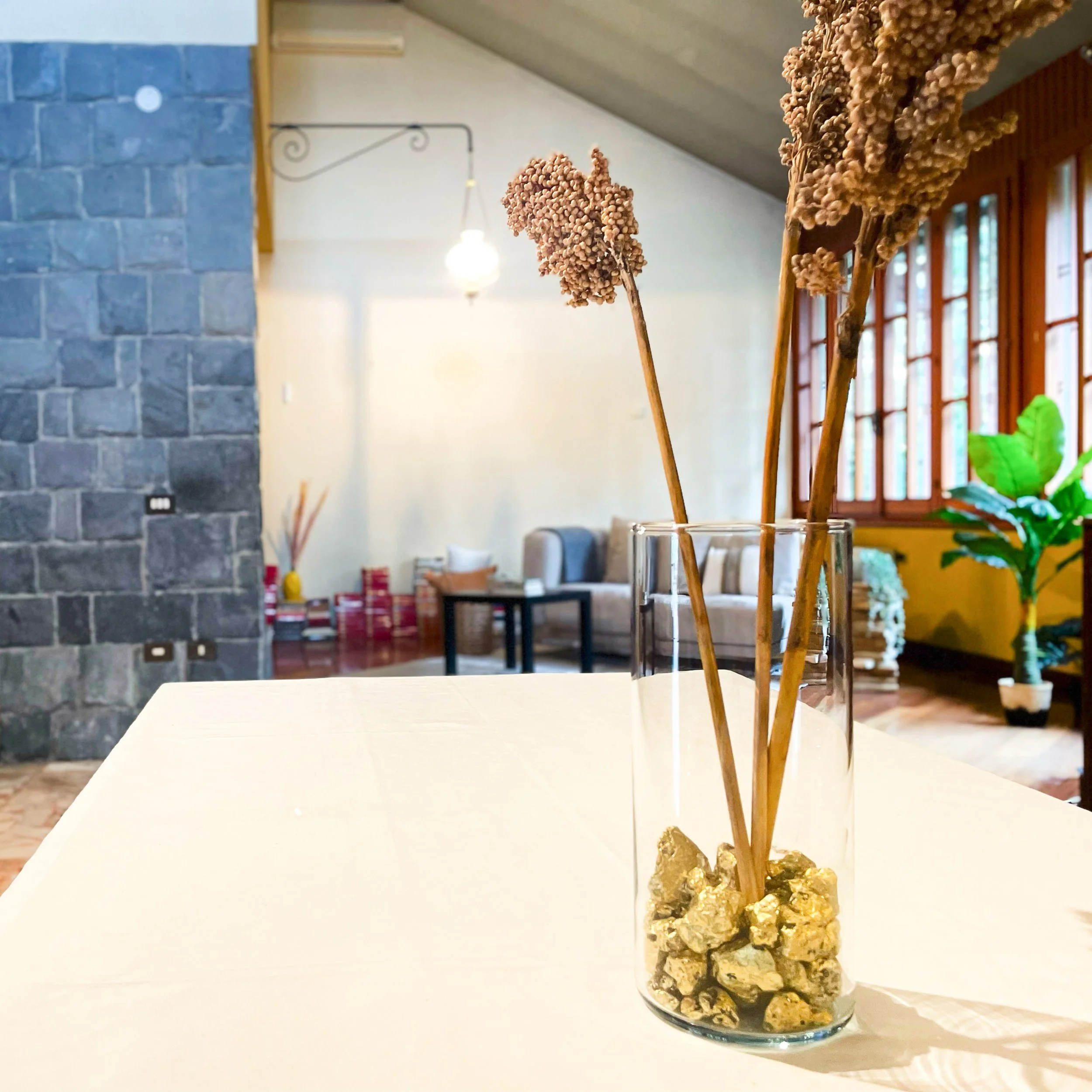 Vaso di vetro su un tavolo con rami secchi e pietre dorate, in uno spazio interno con una parete di pietra e arredi moderni in sottofondo.