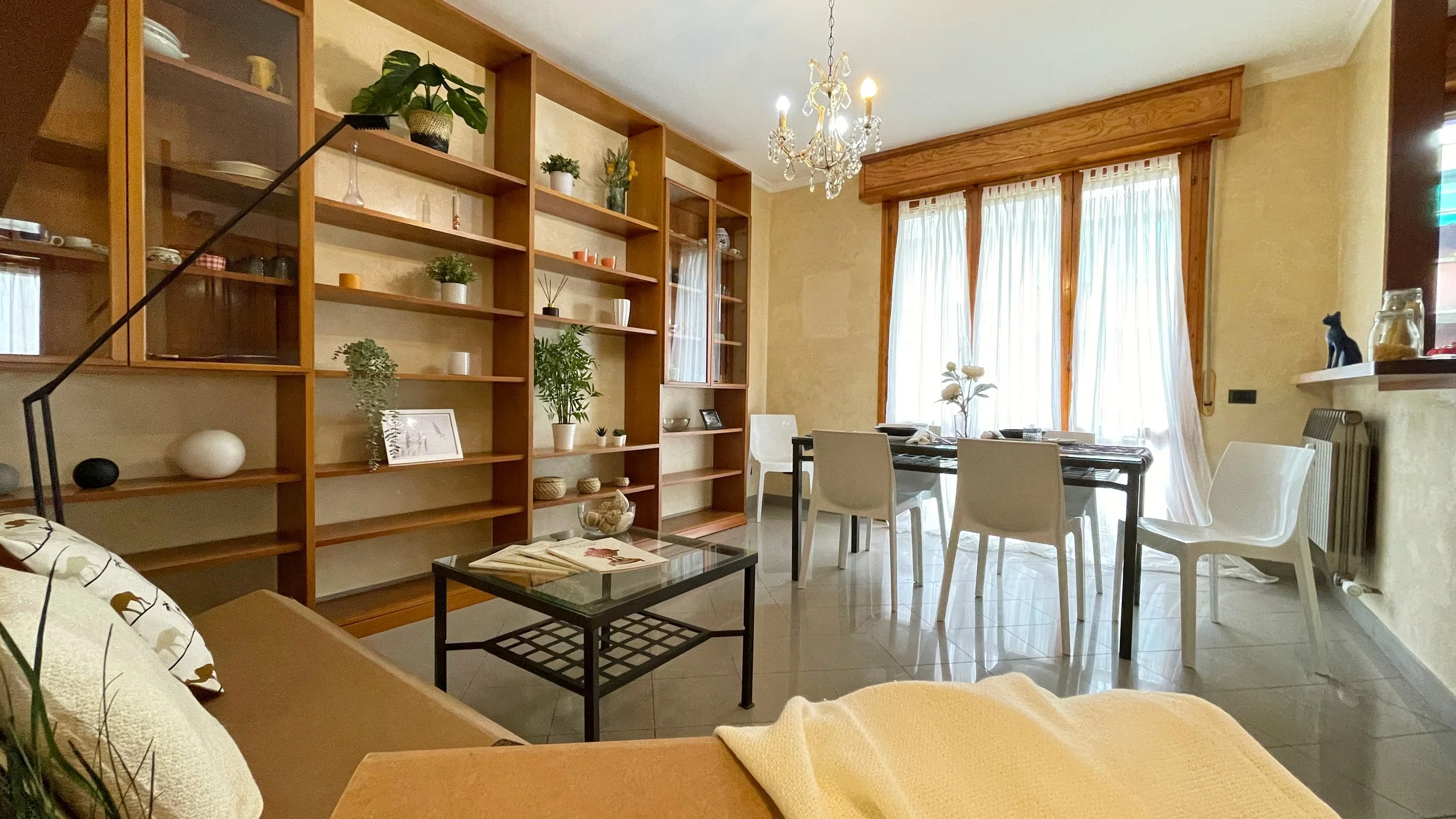 Soggiorno luminoso con scaffali in legno, tavolo da pranzo con sedie bianche, lampadario decorativo e ampie finestre con tende bianche trasparenti.