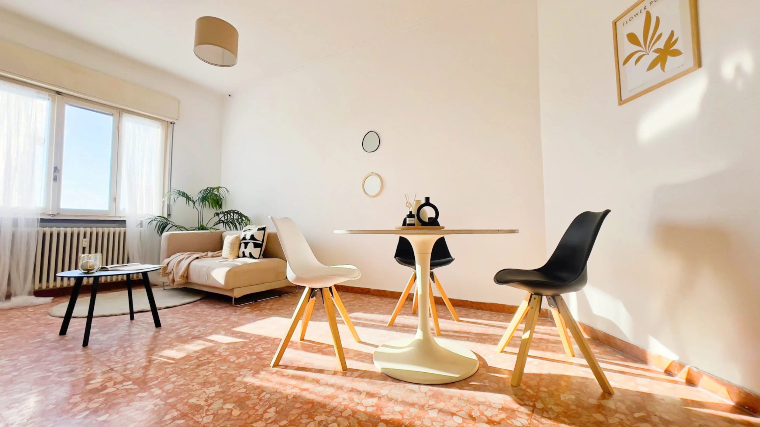 Stanza luminosa con divano beige, tavolo rotondo bianco, sedie moderne, pianta, finestra grande, e decorazioni minimaliste sulla parete.