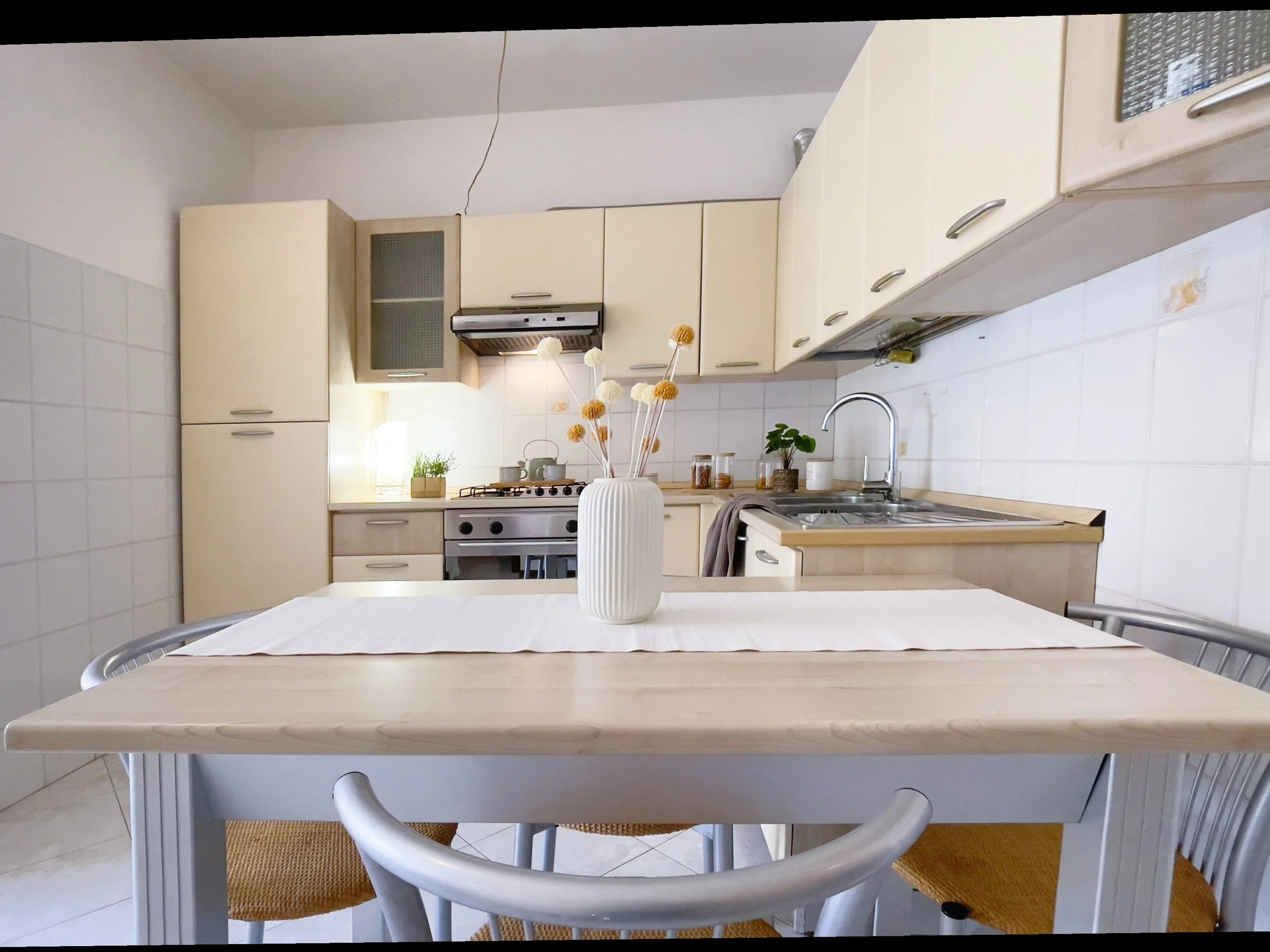 Cucina moderna con arredi in legno chiaro, tavolo con vaso di fiori secchi, lavello in acciaio inox, e stoviglie sulla cucina a gas.