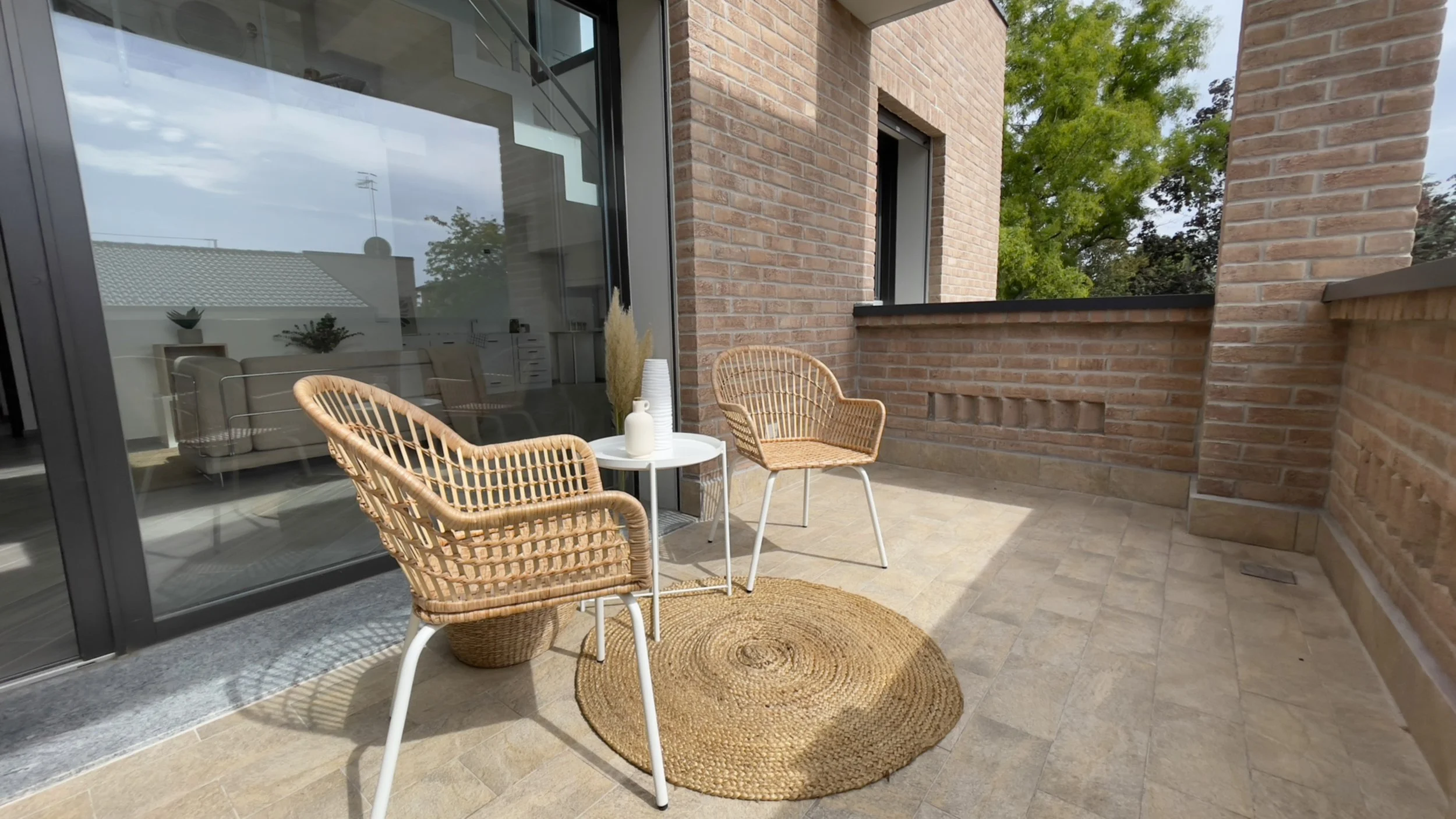 Terrazza moderna con due sedie in rattan e un tavolino bianco, pavimento in piastrelle beige, muri di mattoni, e vista di alberi verdi.