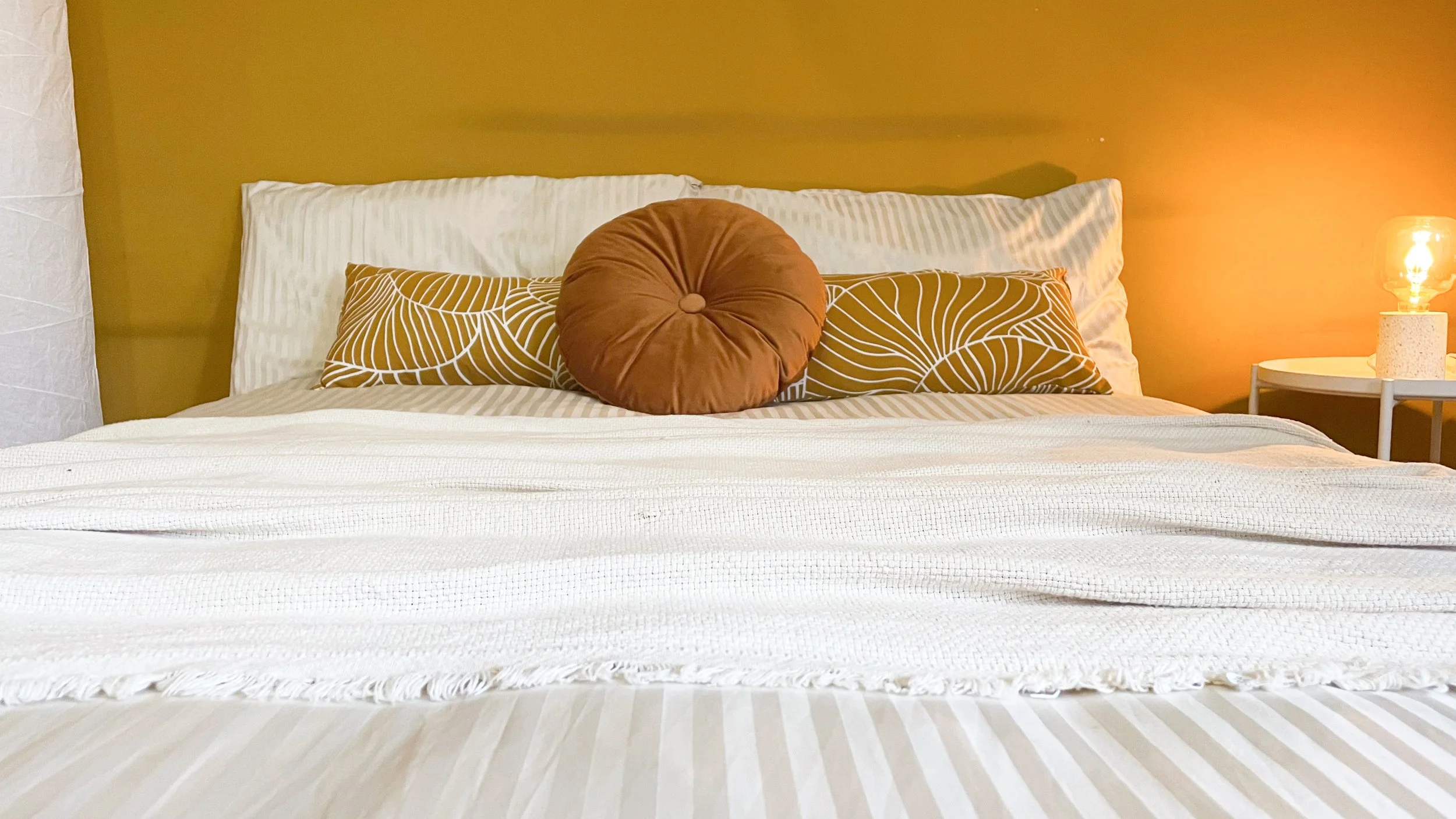 Camera da letto con letto matrimoniale, cuscini decorativi arancioni e gialli, coperta bianca, parete color ocra e lampada accesa su un comodino bianco.