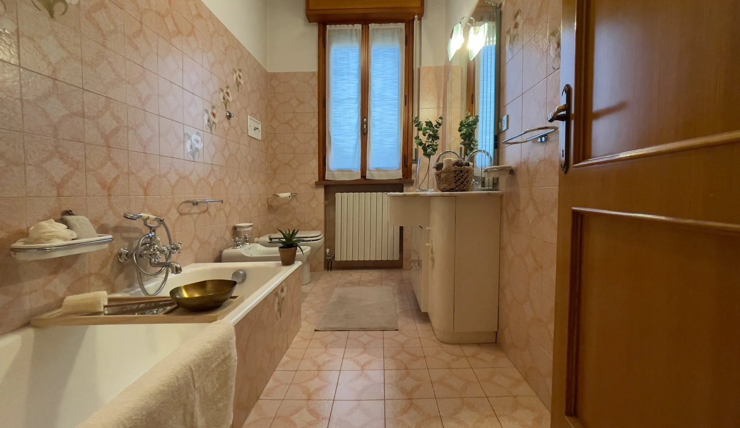 Bagno con vasca da bagno, lavandino, specchio, finestra, piastrelle rosa e decorazioni vegetali.