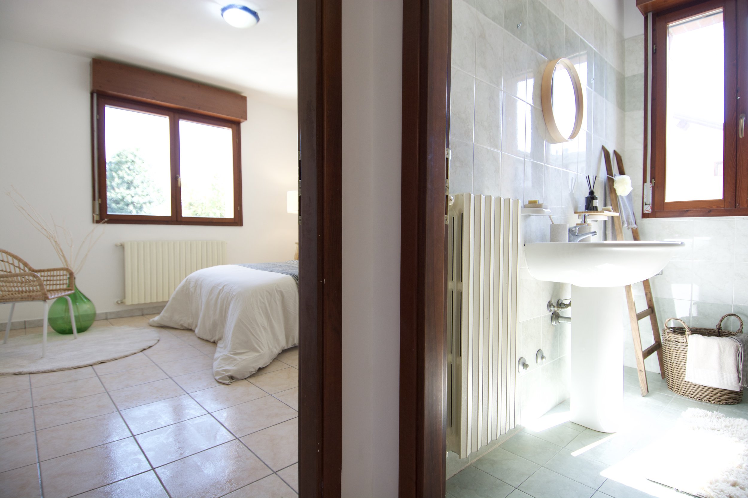 Interno di una casa con una vista su due stanze: una camera da letto con letto, finestra e sedia in vimini a sinistra, e un bagno con lavandino, specchio e cesto a destra.