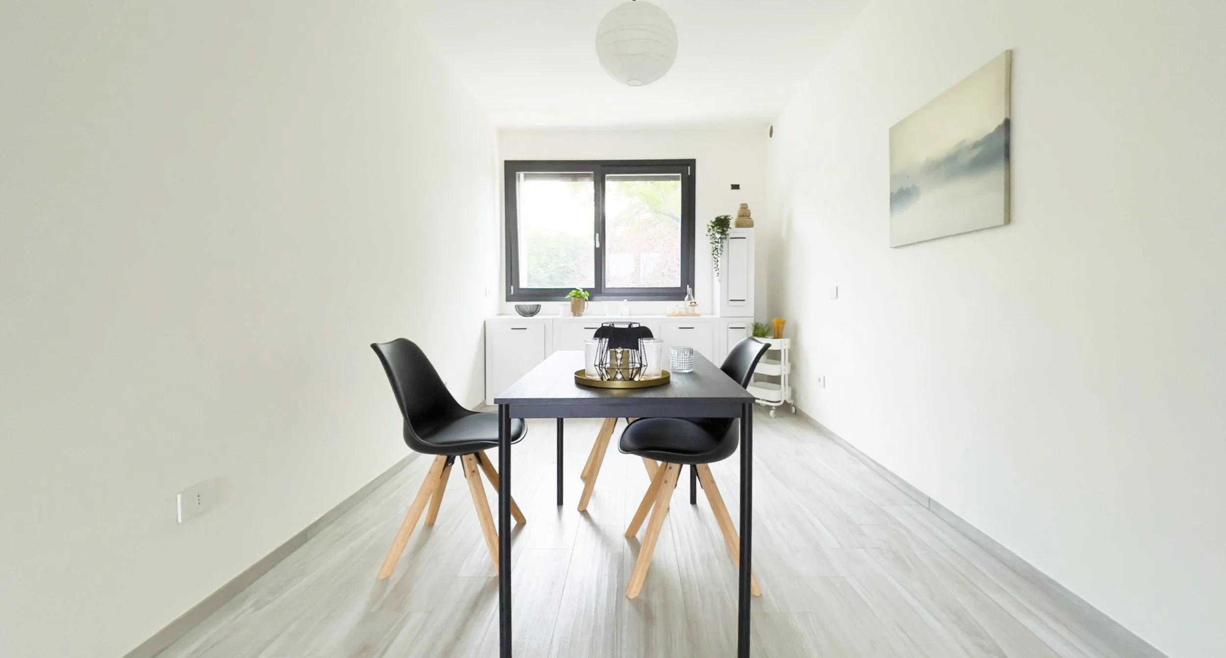 Sala da pranzo moderna con tavolo nero, due sedie nere, pavimento in legno chiaro, finestra grande, decorazione semplice e lampada a sospensione bianca.