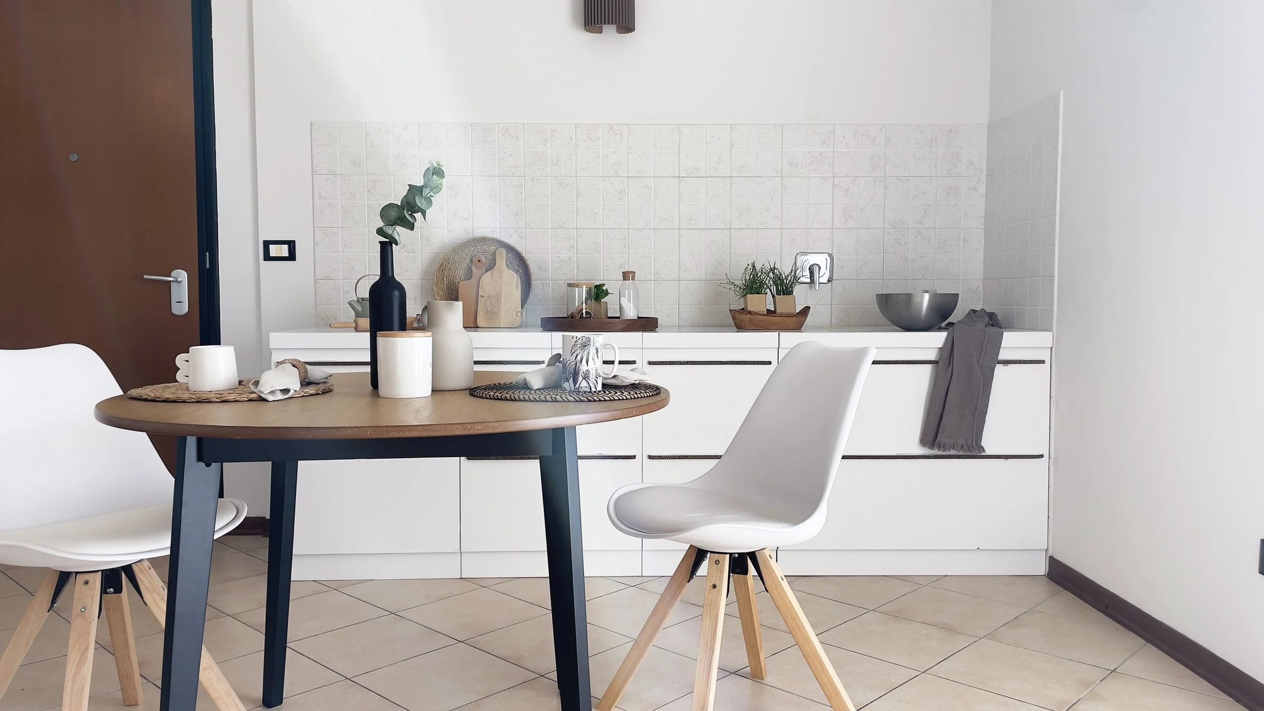 Cucina moderna con tavolo rotondo e sedie bianche, decorazioni minimaliste, parete piastrellata chiara, e accessori come tazze e vasi.