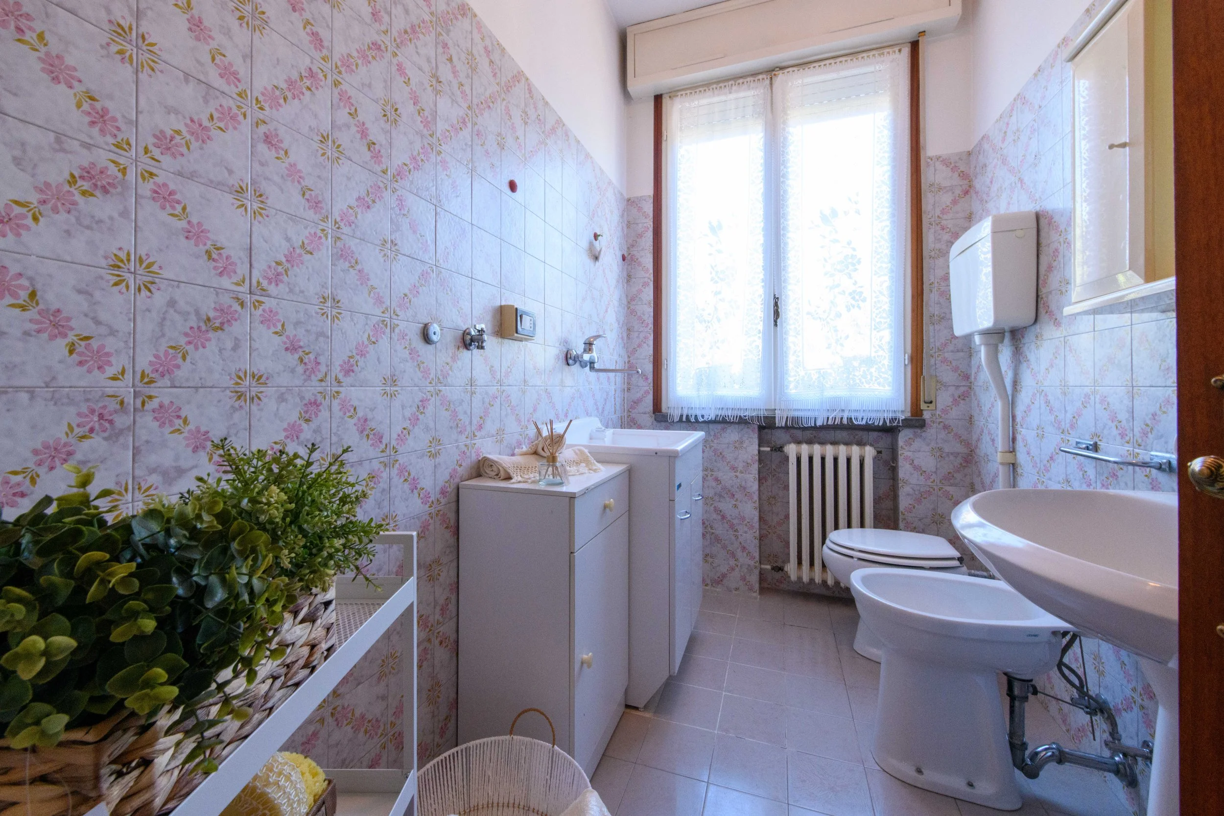 Bagno con pareti decorate con piastrelle floreali, sanitari bianchi, lavabo, bidet e WC. Finestra con tende, mobile con asciugamani e articoli da toeletta, e pianta decorativa su uno scaffale.