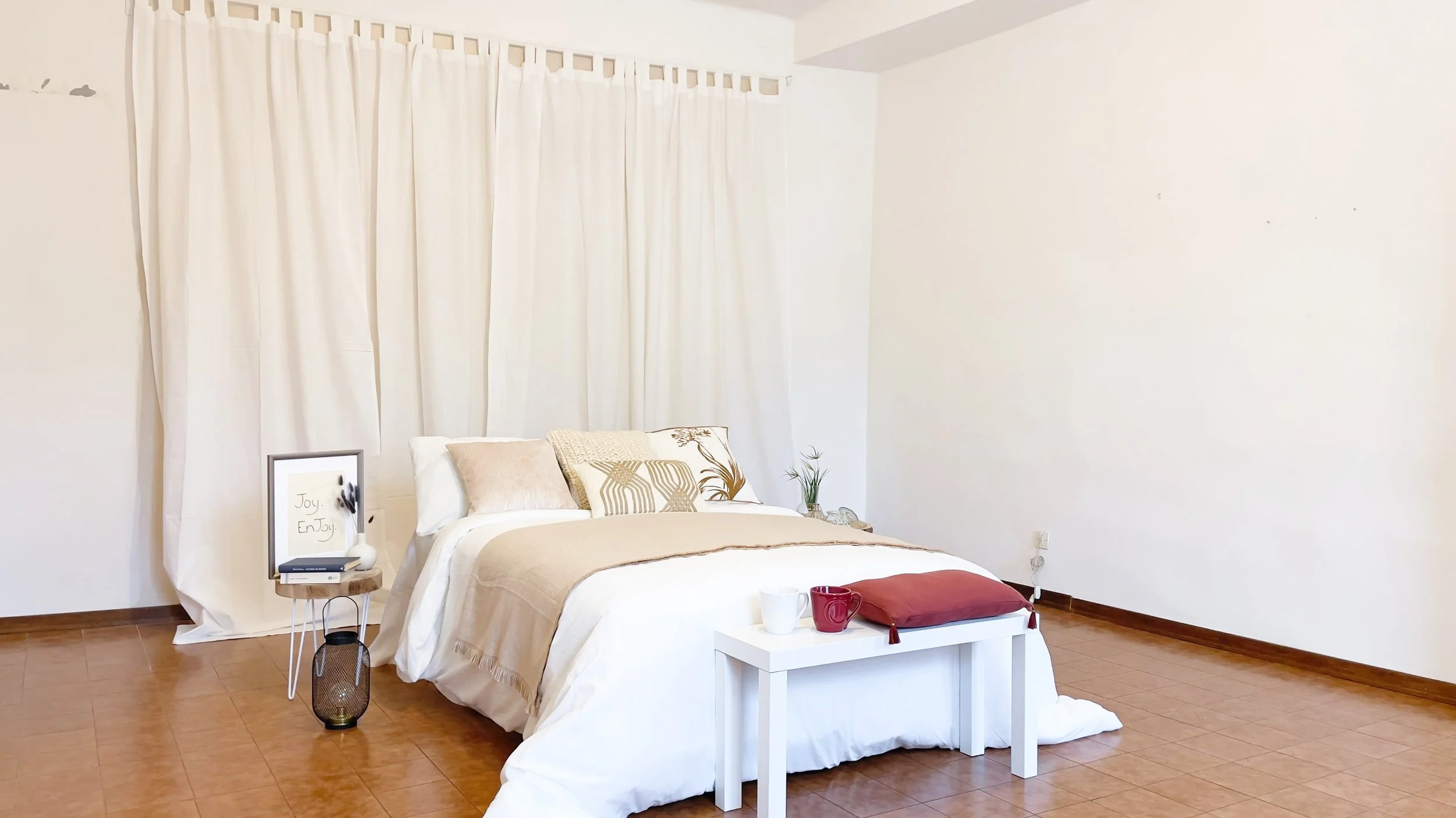 Camera da letto minimalista con letto matrimoniale, cuscini decorativi, tavolino bianco con tazze e cuscino, tende bianche, e parete bianca.