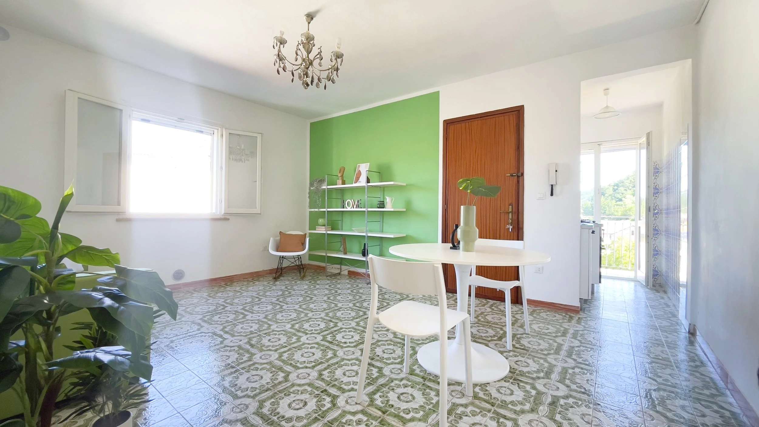 Soggiorno moderno con pareti bianche, una parete verde, pavimento piastrellato con motivi geometrici verdi e bianchi. C'è un tavolo rotondo bianco con una sedia, una libreria con decorazioni e una finestra che lascia entrare molta luce naturale. Pian
