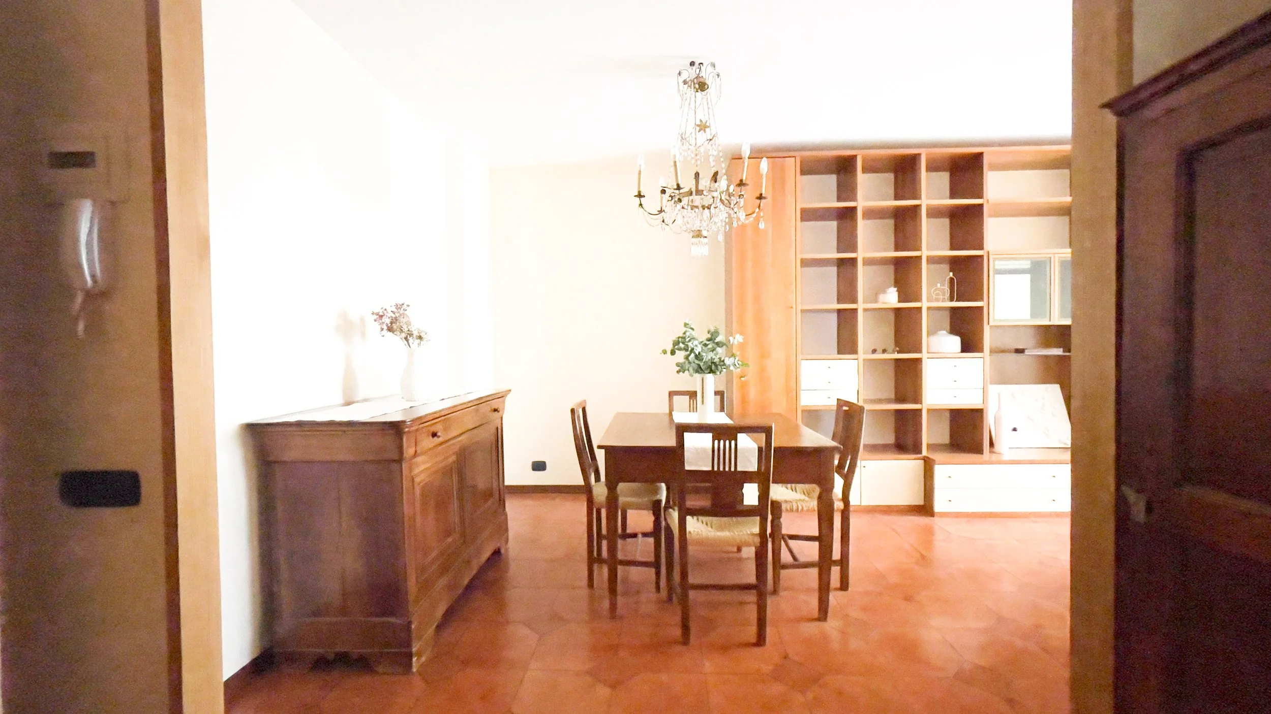 Sala da pranzo con tavolo e sedie in legno, mobile antico, scaffalatura, lampadario e decorazioni floreali. Pavimento in piastrelle rosse.