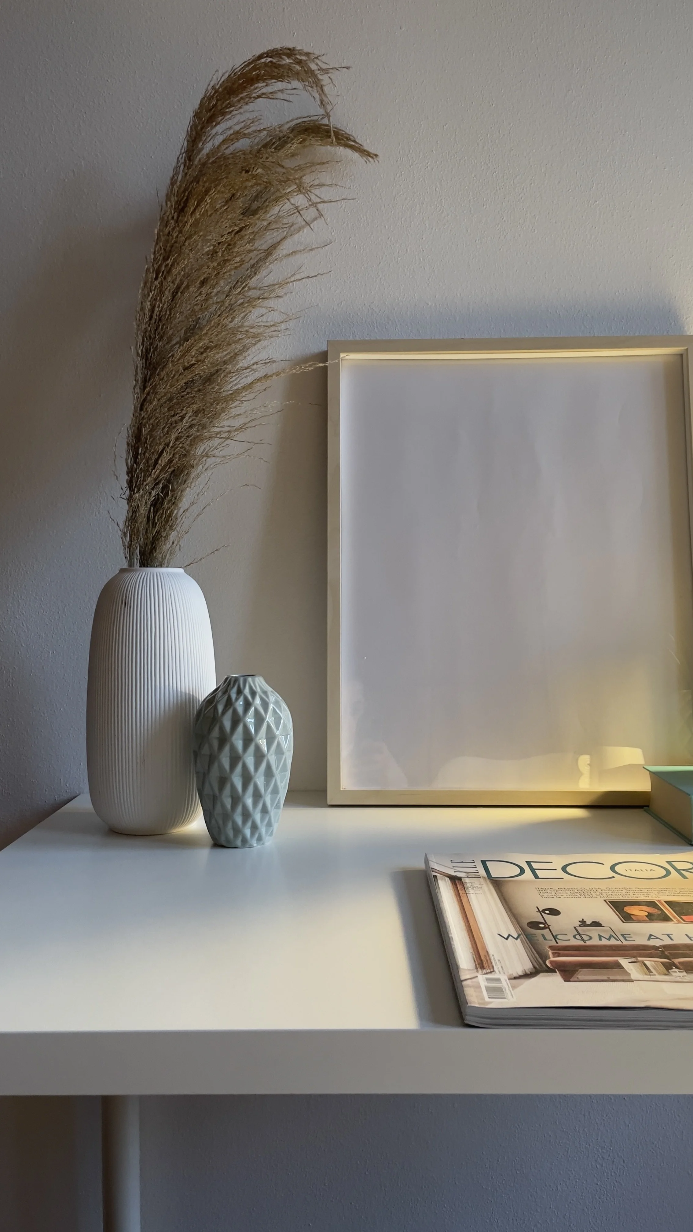 Tavolino bianco con vaso bianco alto con erba secca, vaso blu in ceramica, cornice vuota e rivista di arredamento.