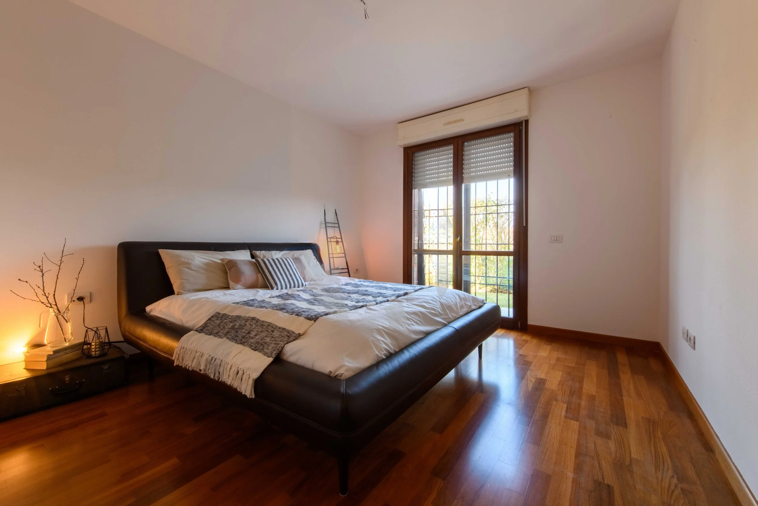 Camera da letto moderna con letto matrimoniale, pavimento in legno, finestra grande con persiane e decorazioni semplici.