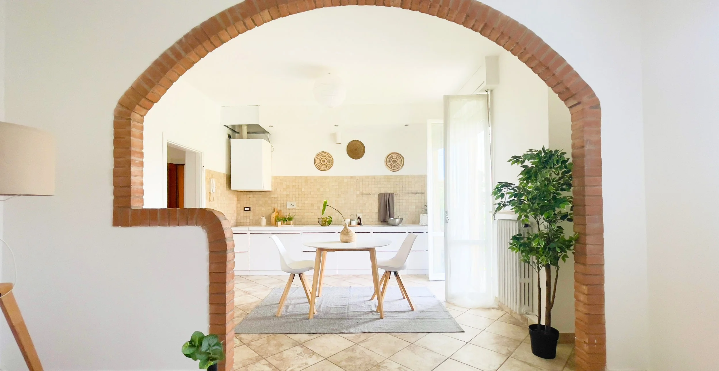 Vista di una cucina moderna con un arco in mattoni. Tavolo rotondo di legno chiaro con sedie bianche su tappeto grigio. Pianta in vaso e decorazioni alle pareti.