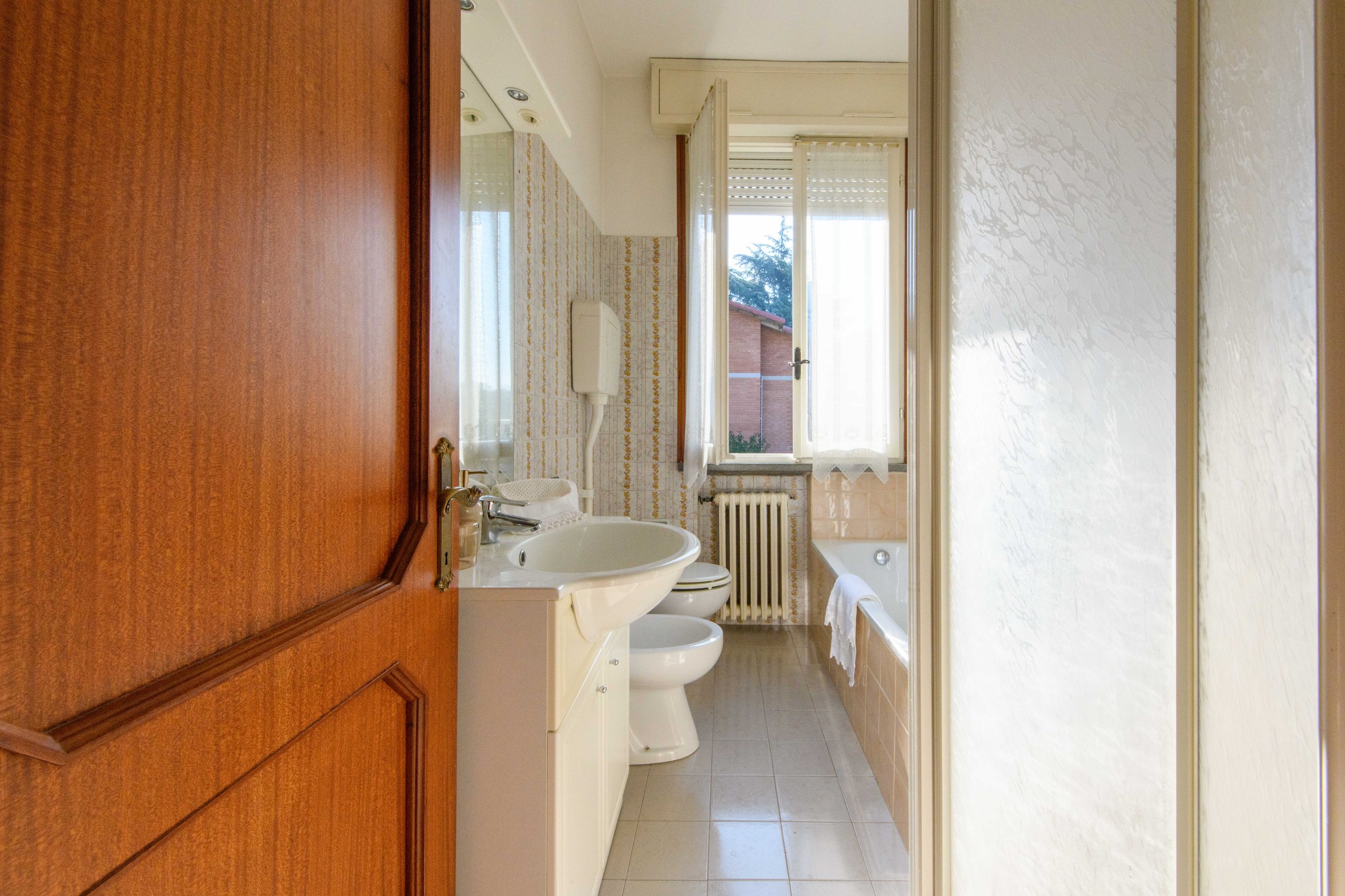 Bagno con piastrelle beige, lavabo, WC, vasca da bagno e finestra con tende bianche.
